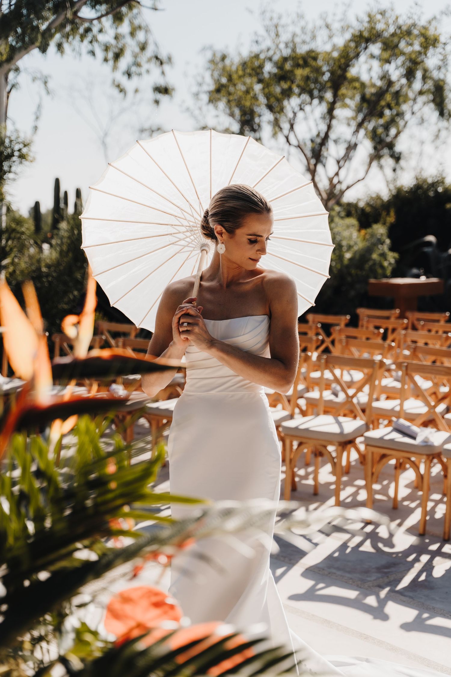 Bride with white parasol poses in outdoor garden setting with wooden chairs.