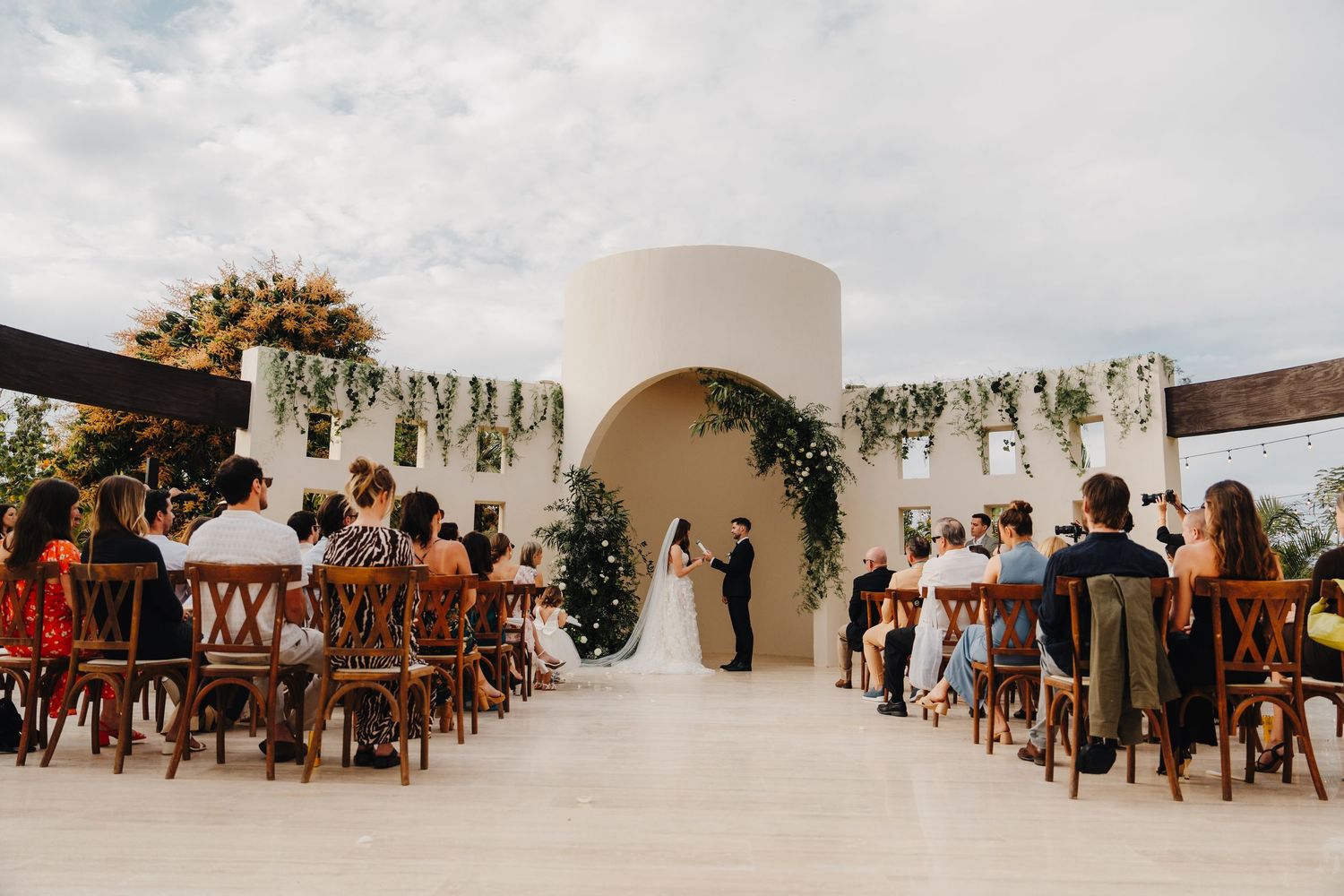 Modern white outdoor wedding ceremony setup with wooden chairs and floral decorations.