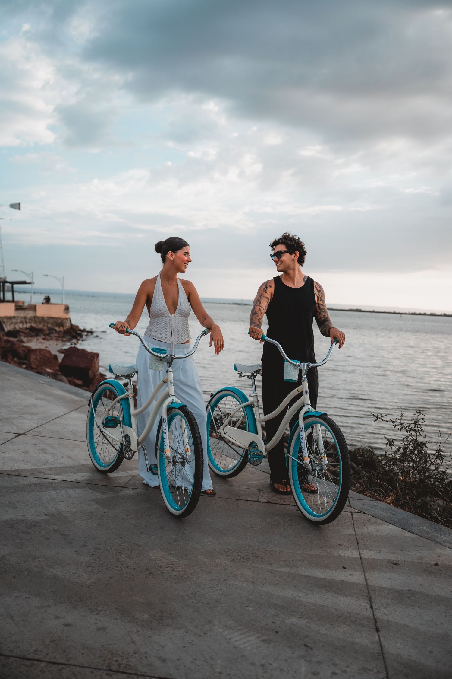 Two people walking turquoise bicycles along waterfront path at dusk.