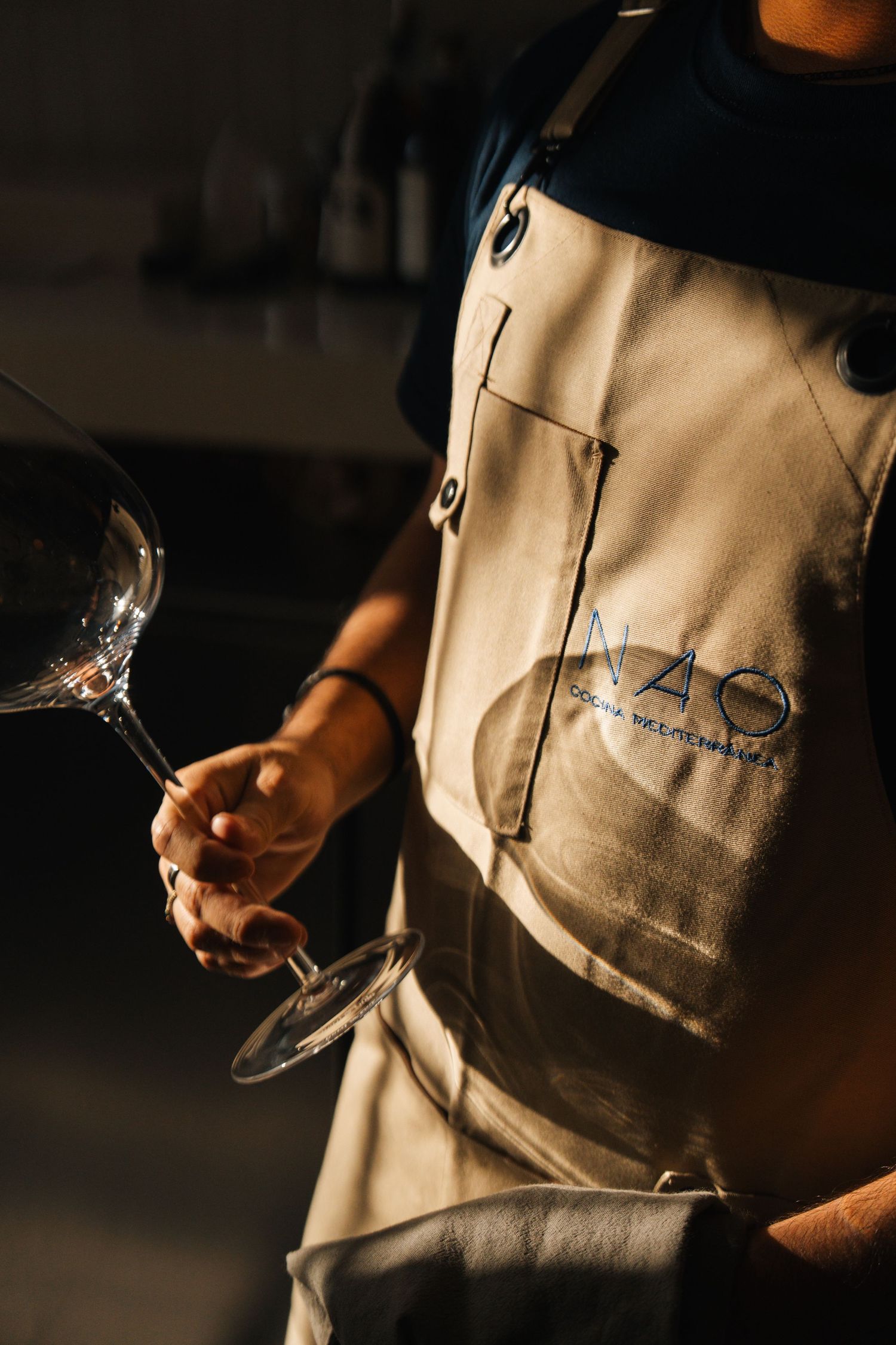 Server wearing tan apron holding wine glass in dimly lit restaurant.