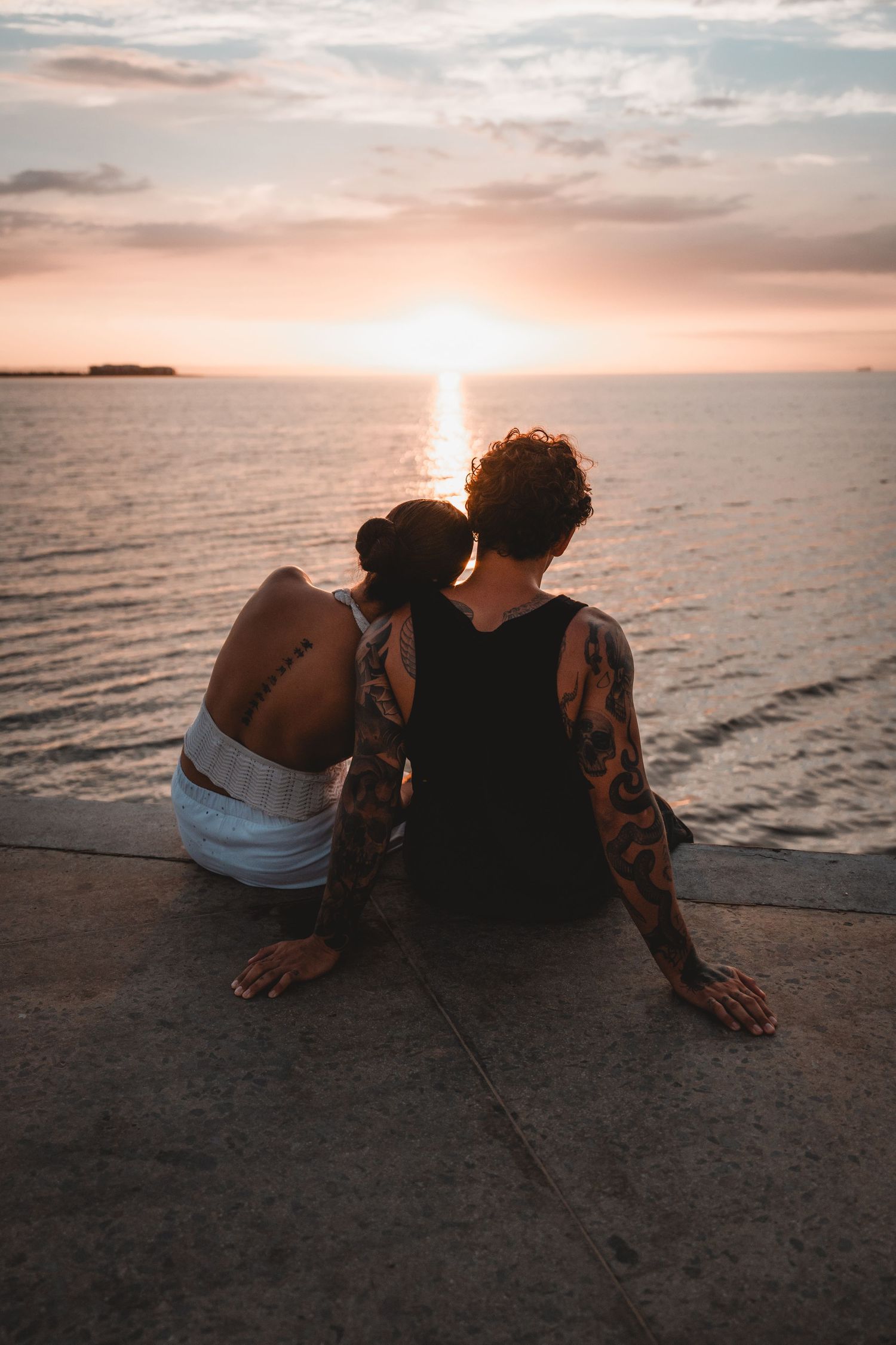 Couple sitting together watching a peaceful ocean sunset from a concrete pier.