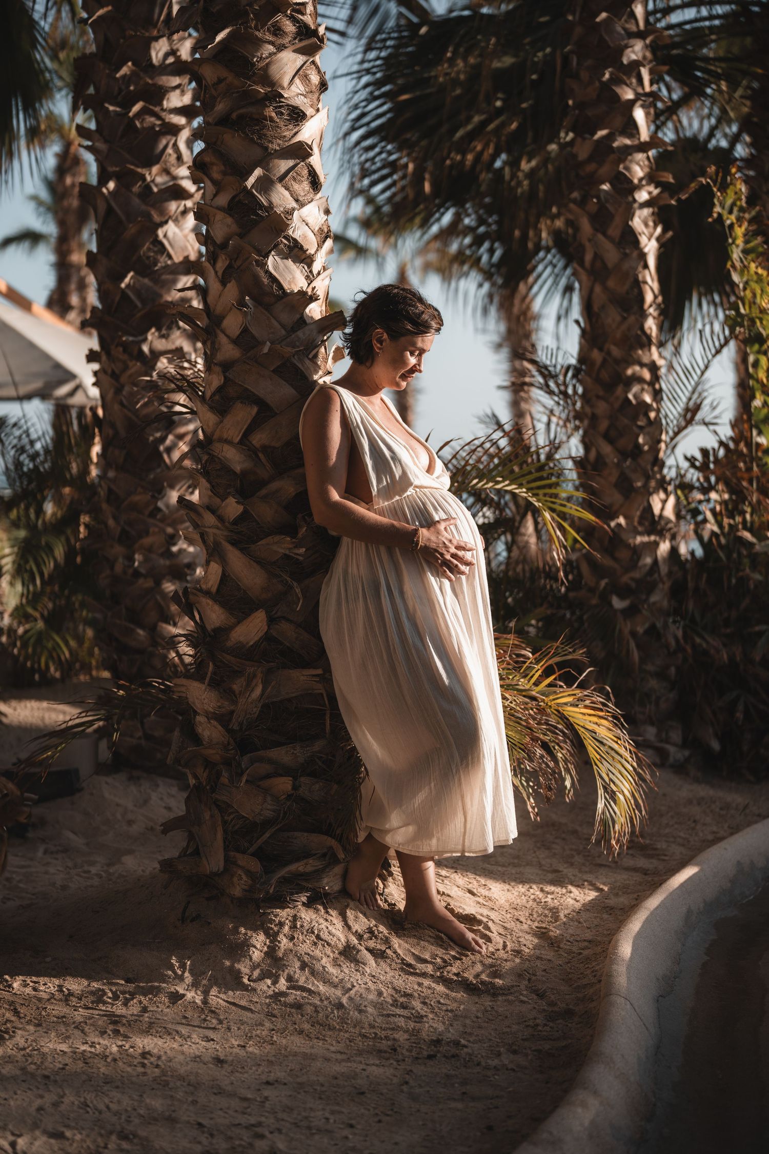 Maternity photoshoot among palm trees at golden hour with flowing white dress.