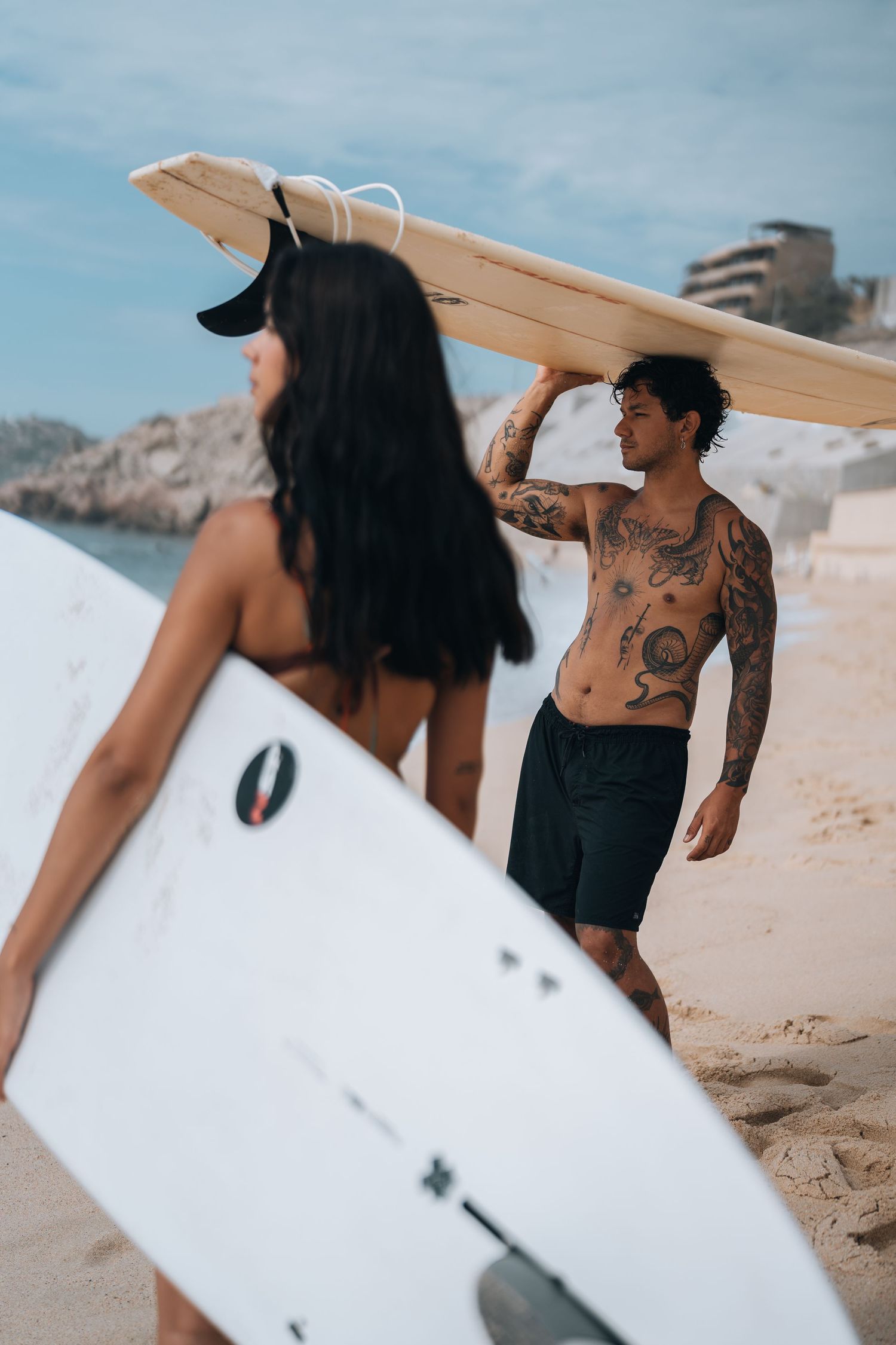 Two surfers walking along sandy beach carrying white surfboards under bright blue sky.
