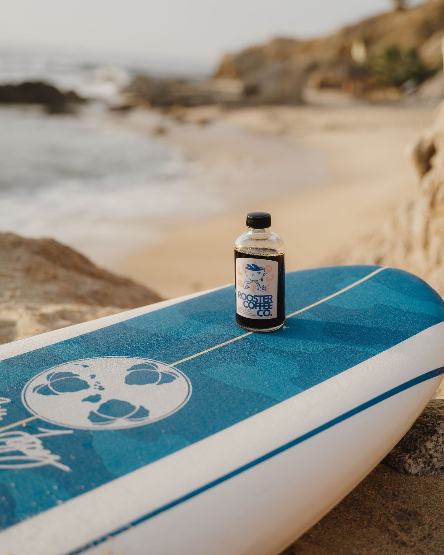 Blue and white surfboard with bottle placed on beach rocks.