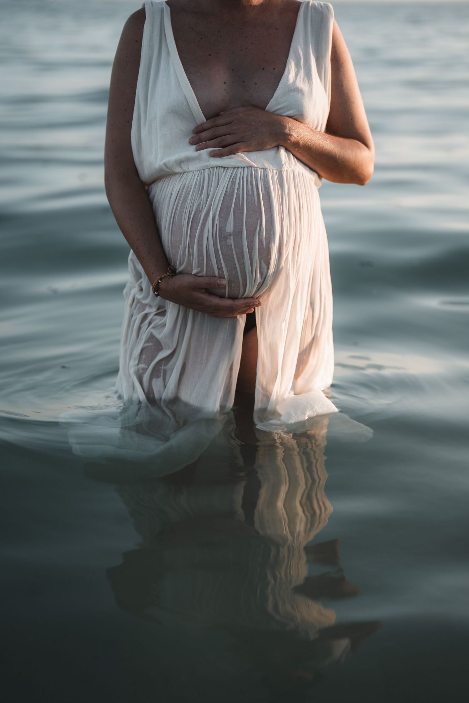 Ethereal maternity photo featuring flowing white dress partially submerged in calm ocean waters.