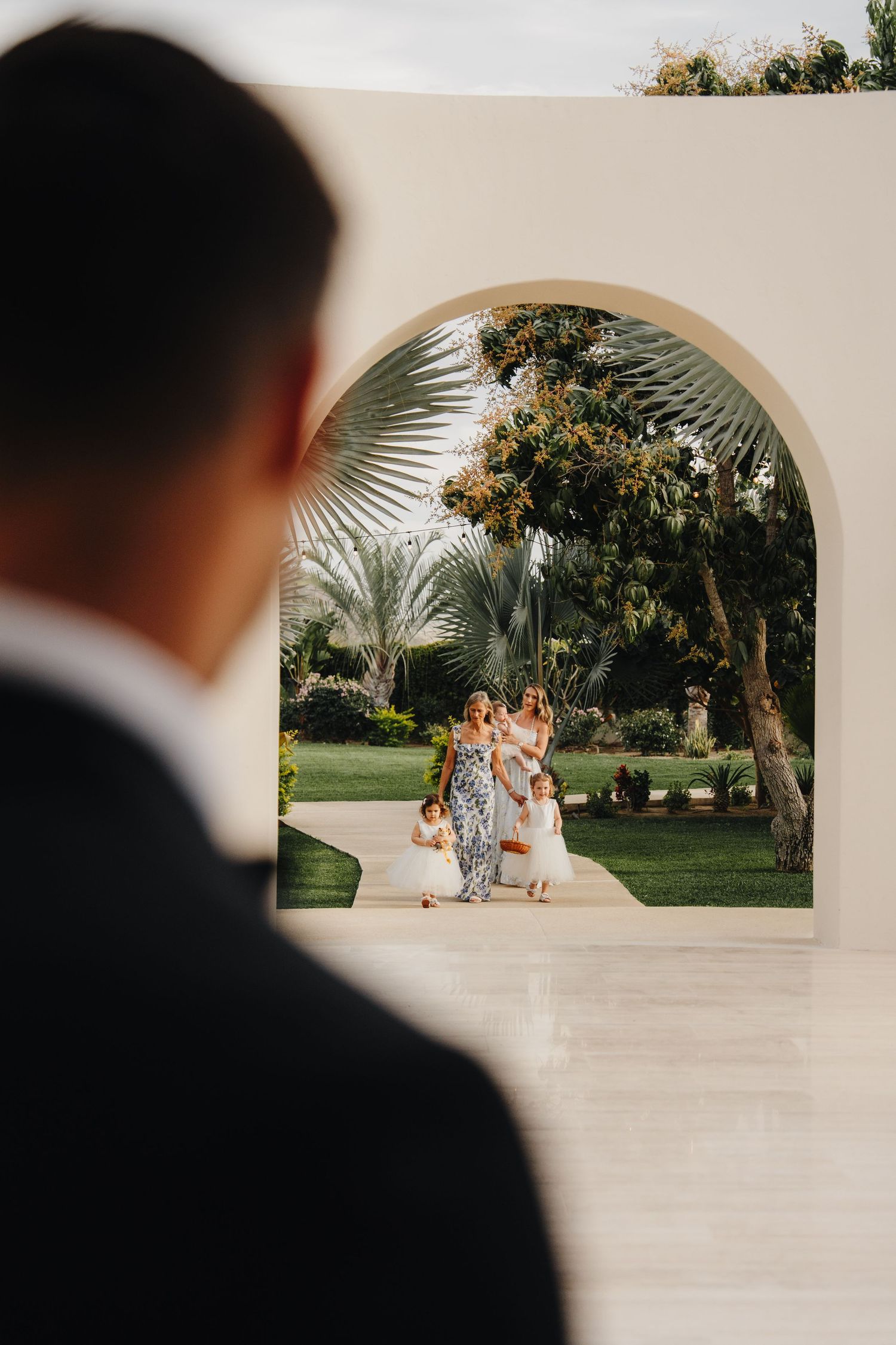 White archway frames an outdoor wedding venue with palm trees and sunlit garden scenery.