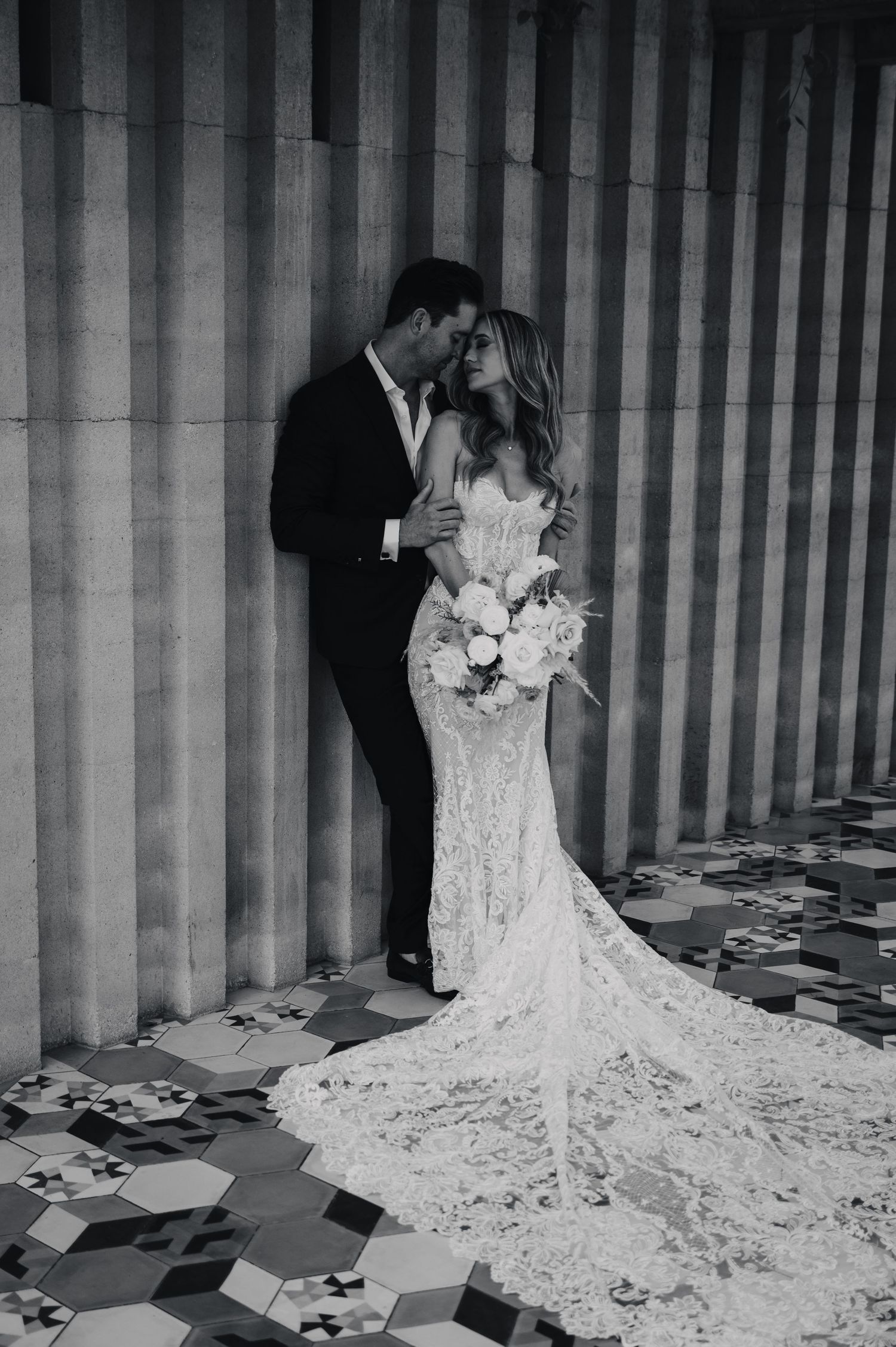 Wedding couple posing against architectural columns with flowing lace dress trail.
