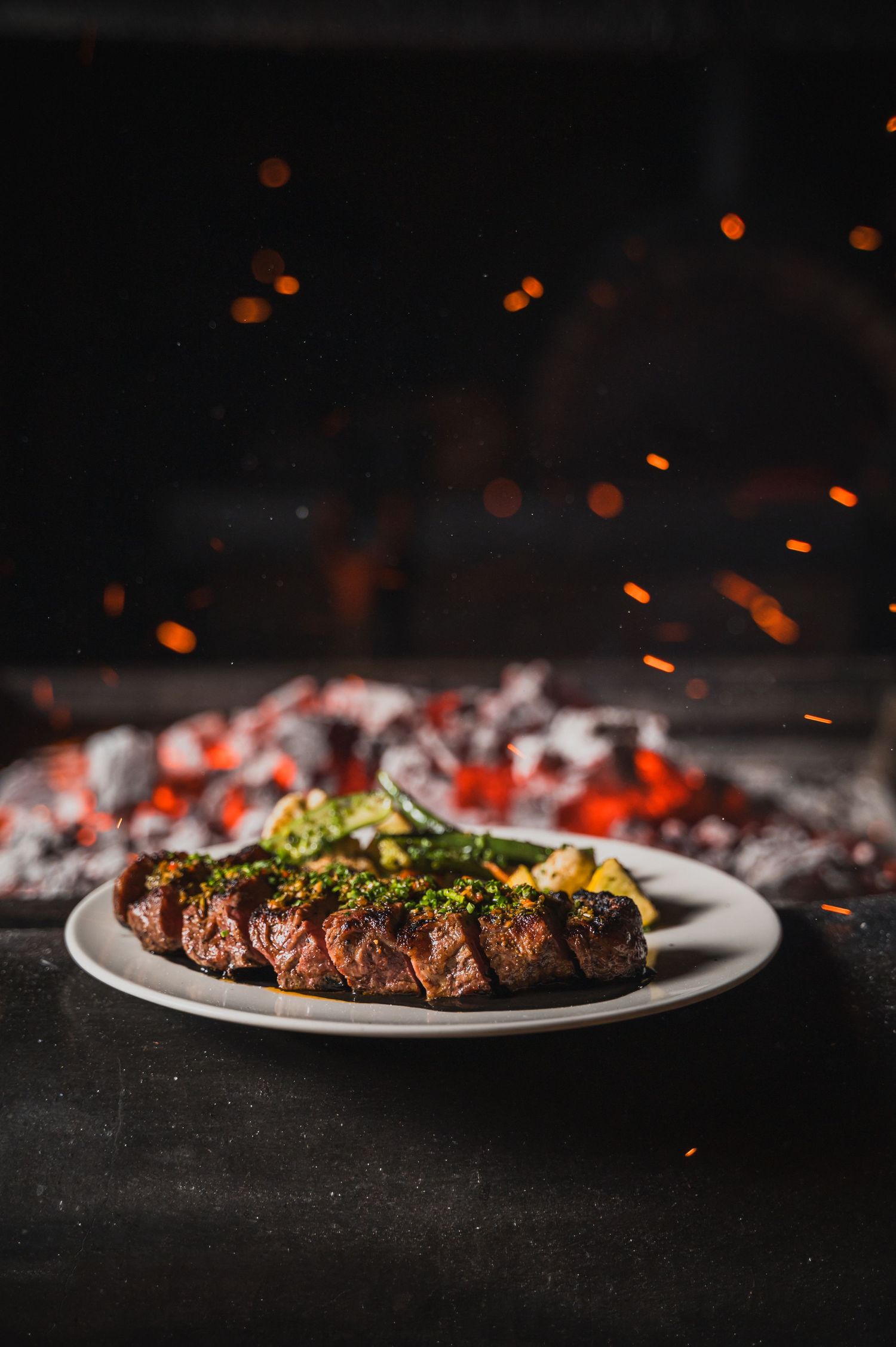 Grilled steak on a white plate with charred vegetables and ember sparks in dark lighting.