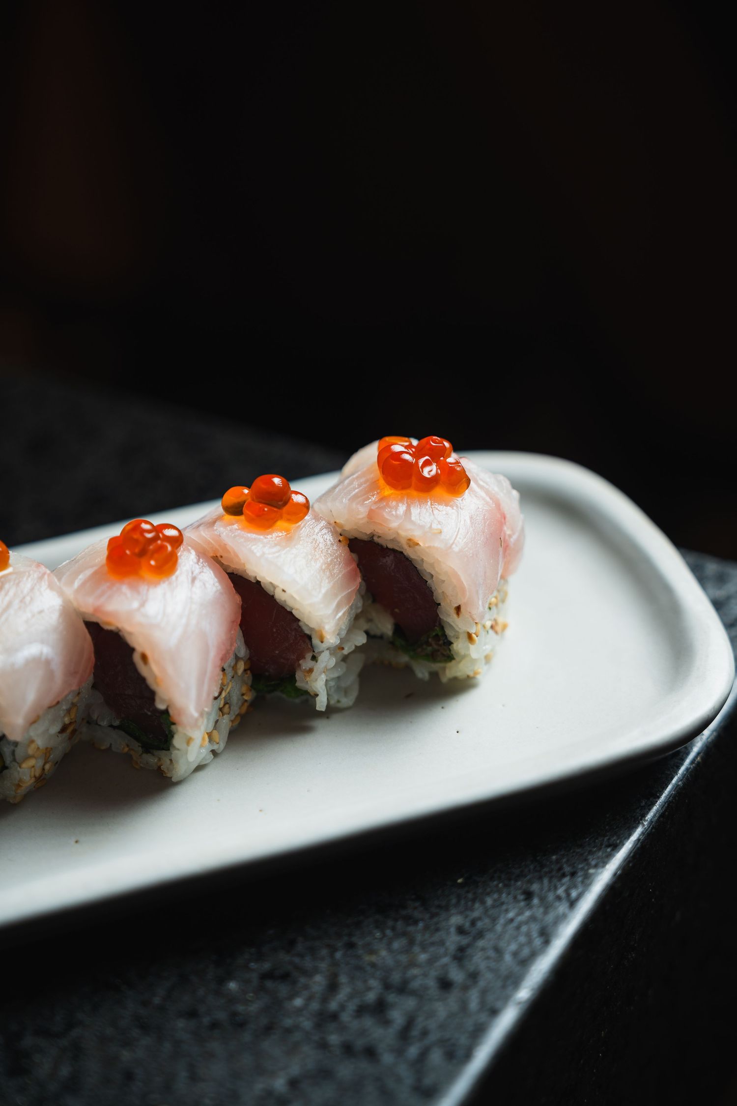 Fresh sushi rolls topped with raw fish and bright orange salmon roe caviar on a white ceramic plate.