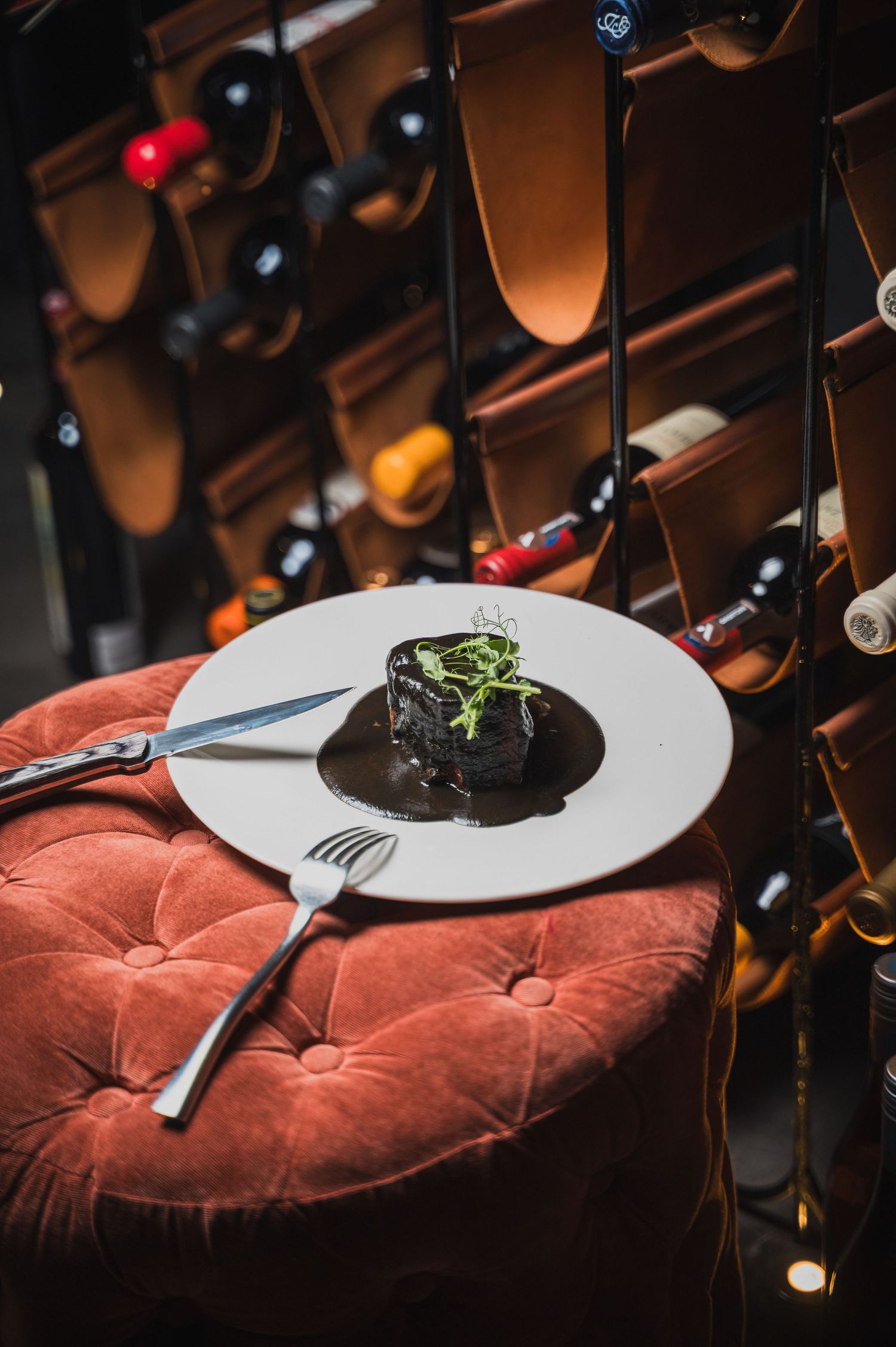 Elegant plated dish on white ceramic plate against wine rack background with red velvet cushion.