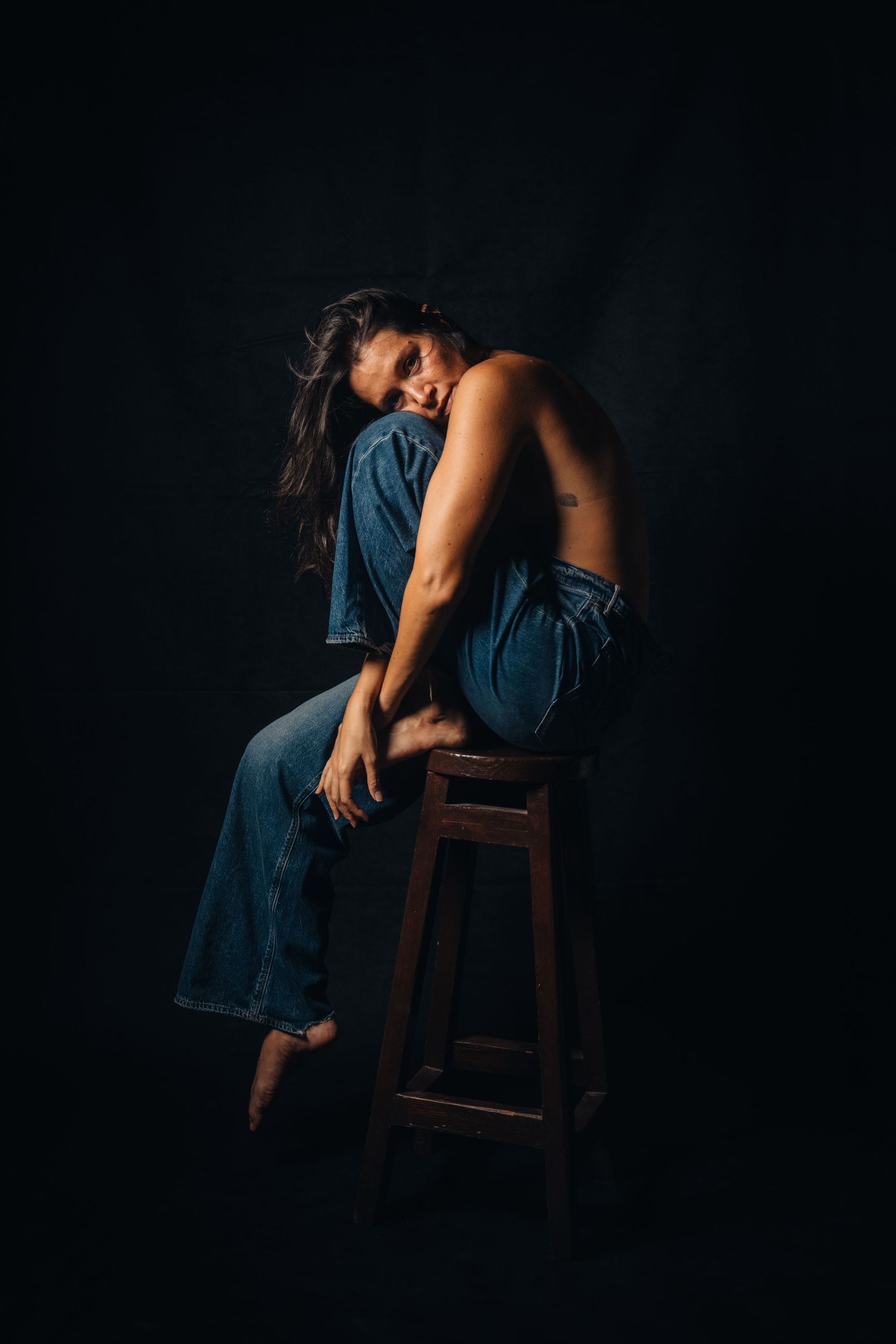Artistic low-key photograph of a figure in blue denim leaning over a wooden stool.