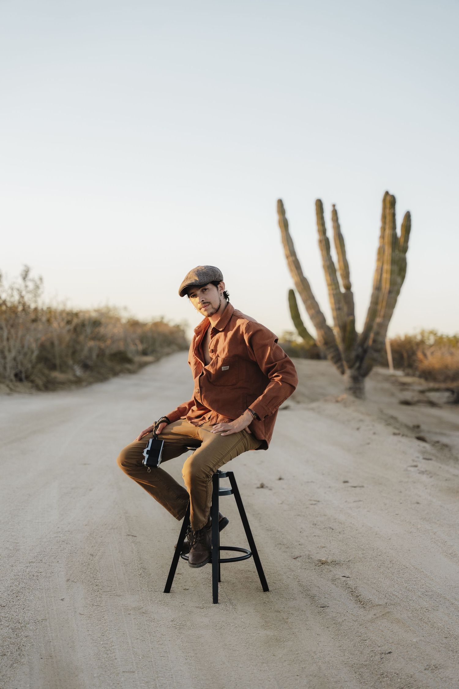 Figure seated on stool on dirt road with saguaro cactus in desert landscape at sunset.