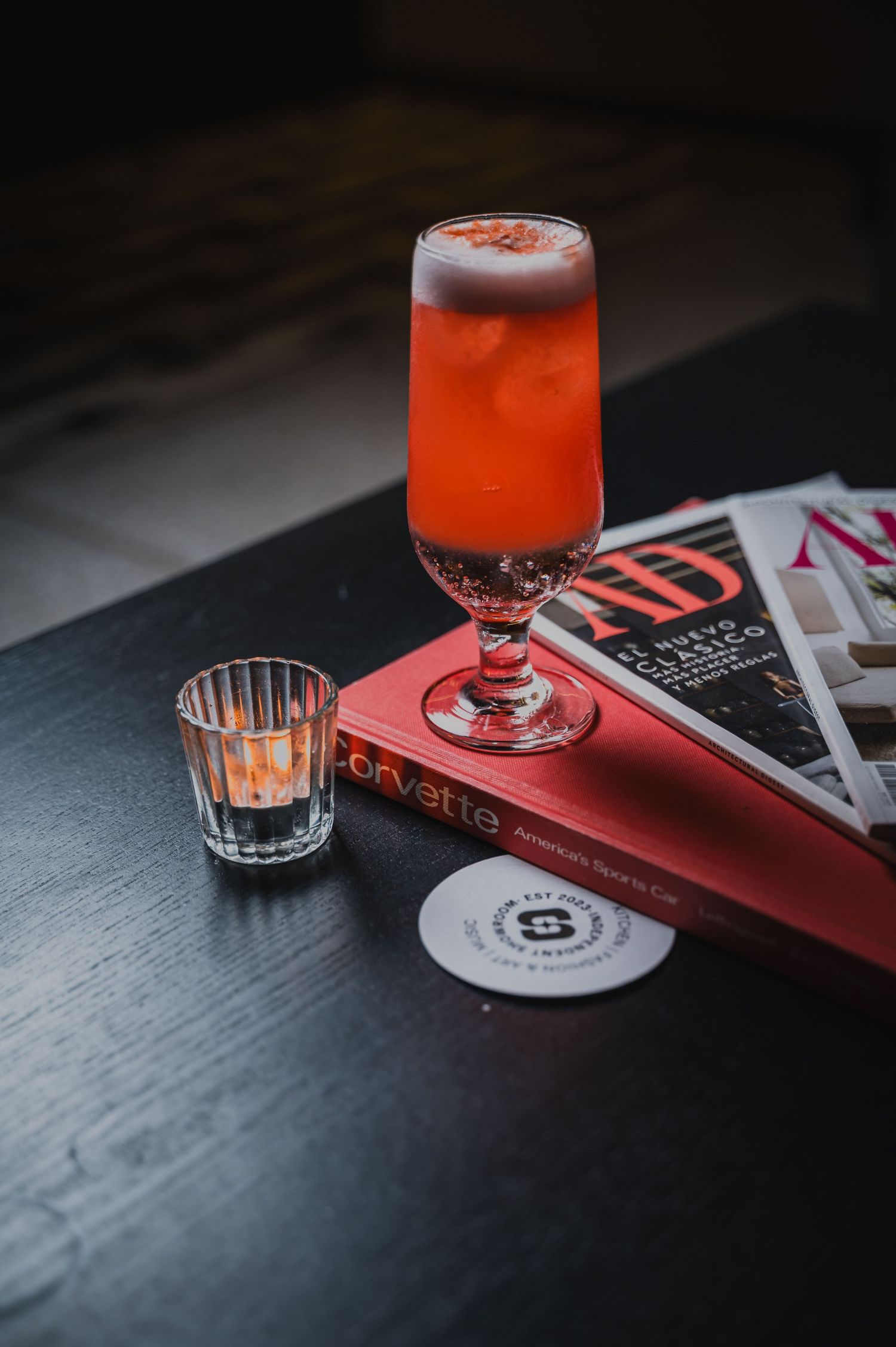Red cocktail in vintage glassware with bar accessories and coasters on dark surface.