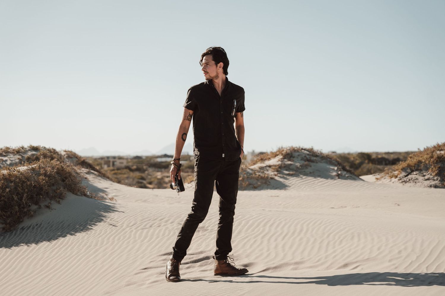 Person in all black outfit stands on sandy dunes against clear sky.