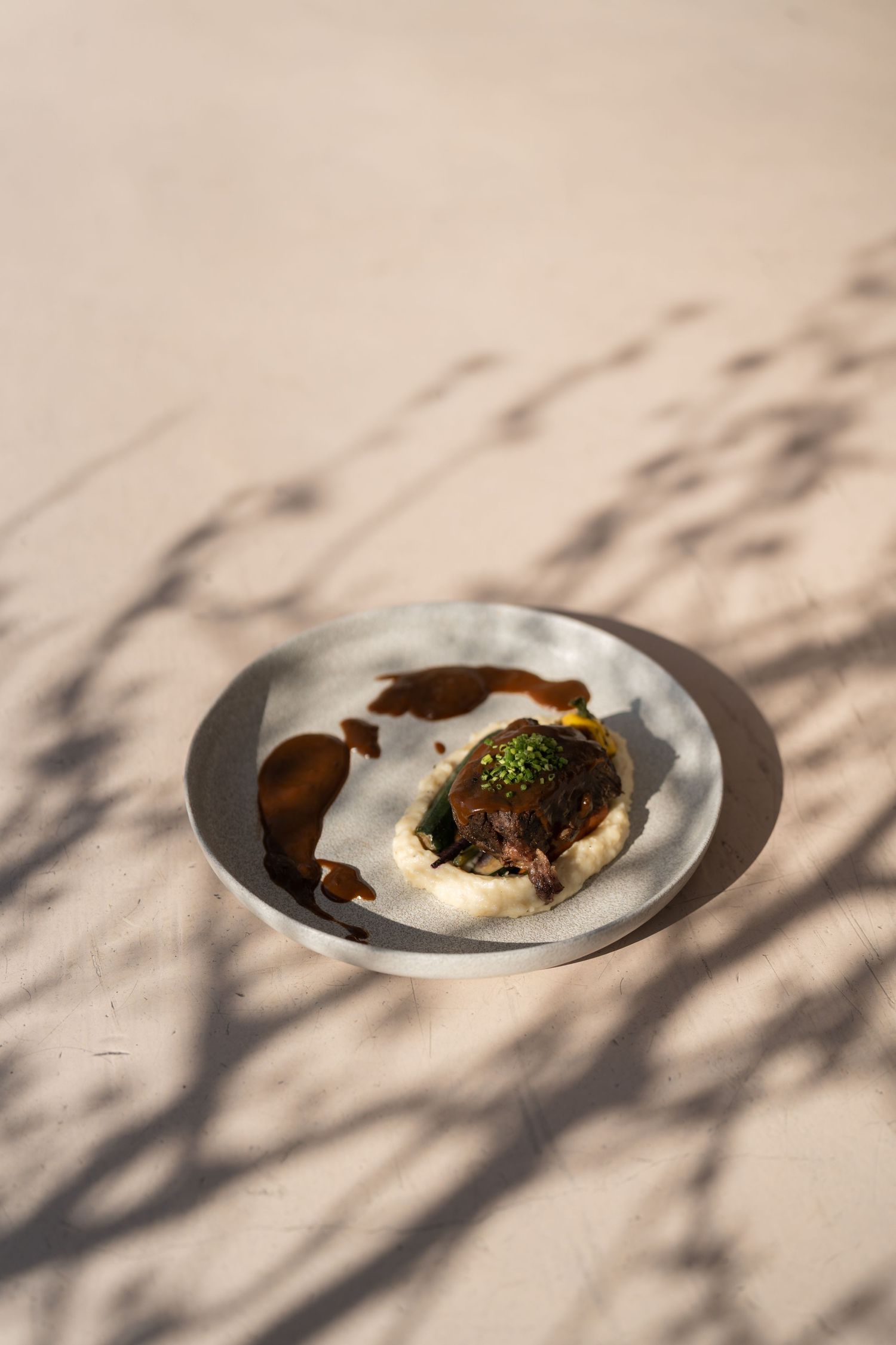Plated oyster dish with garnish and sauce on metal plate under dappled sunlight.