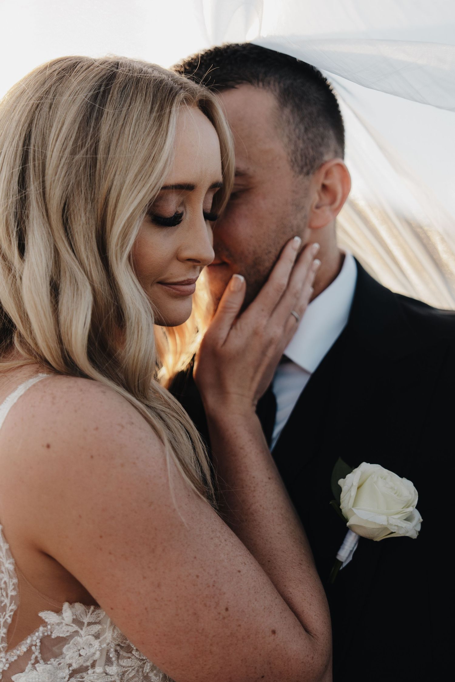 Intimate moment between newlyweds under sunlit veil with white rose boutonniere.