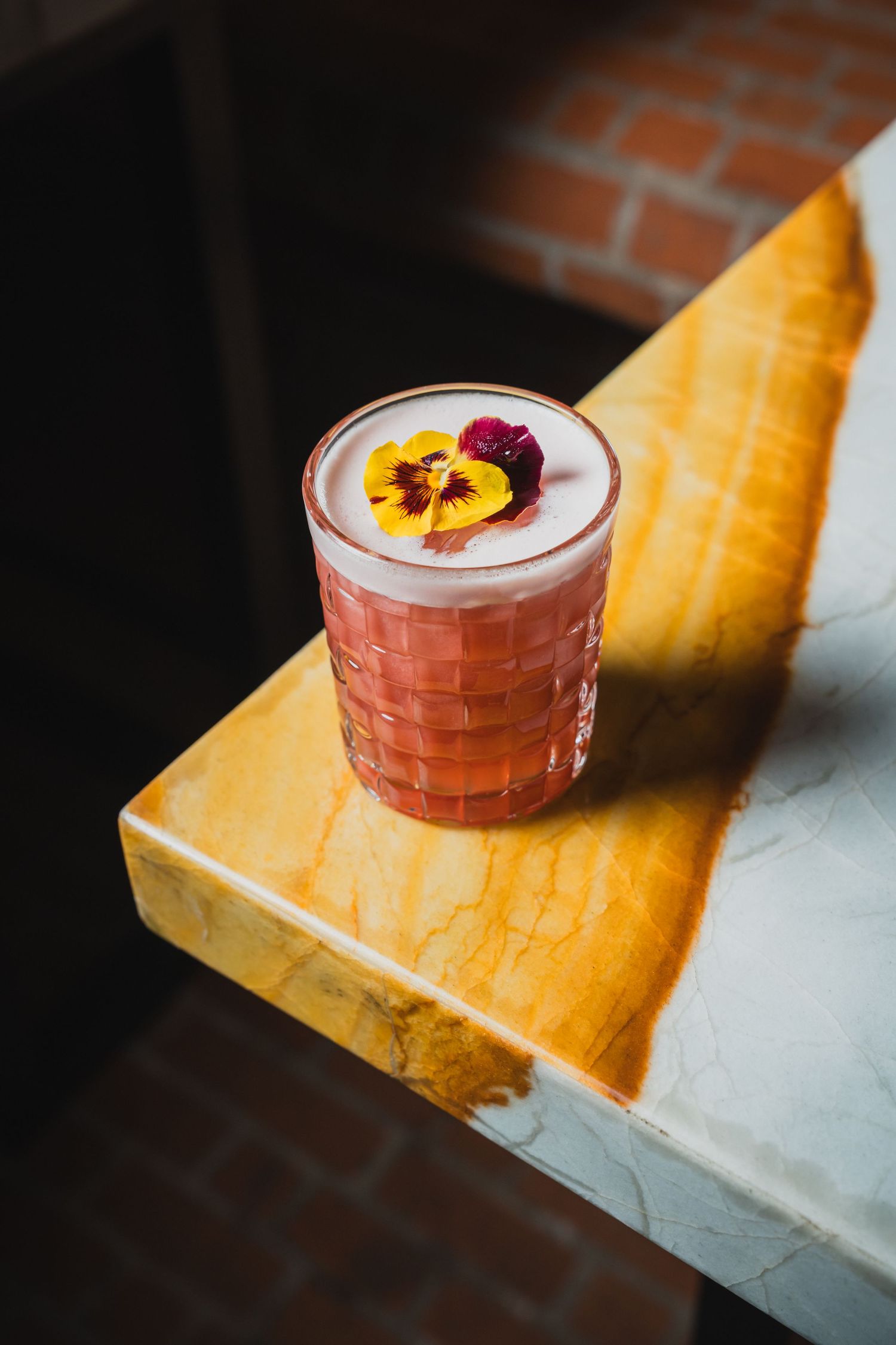 Pink cocktail garnished with fruit served in a crystal glass on a marble surface.