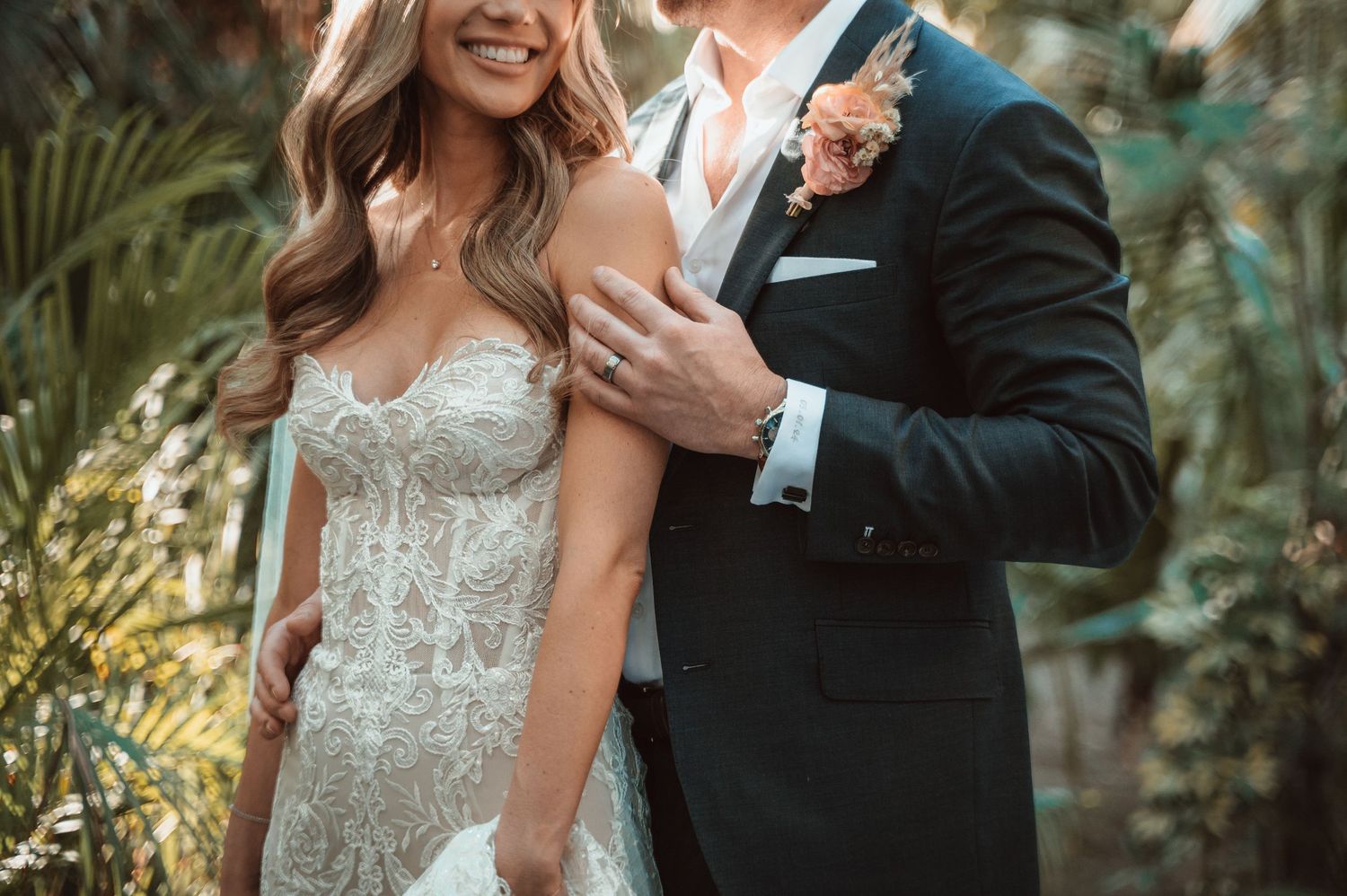 Couple embracing in formal wedding attire surrounded by tropical greenery.