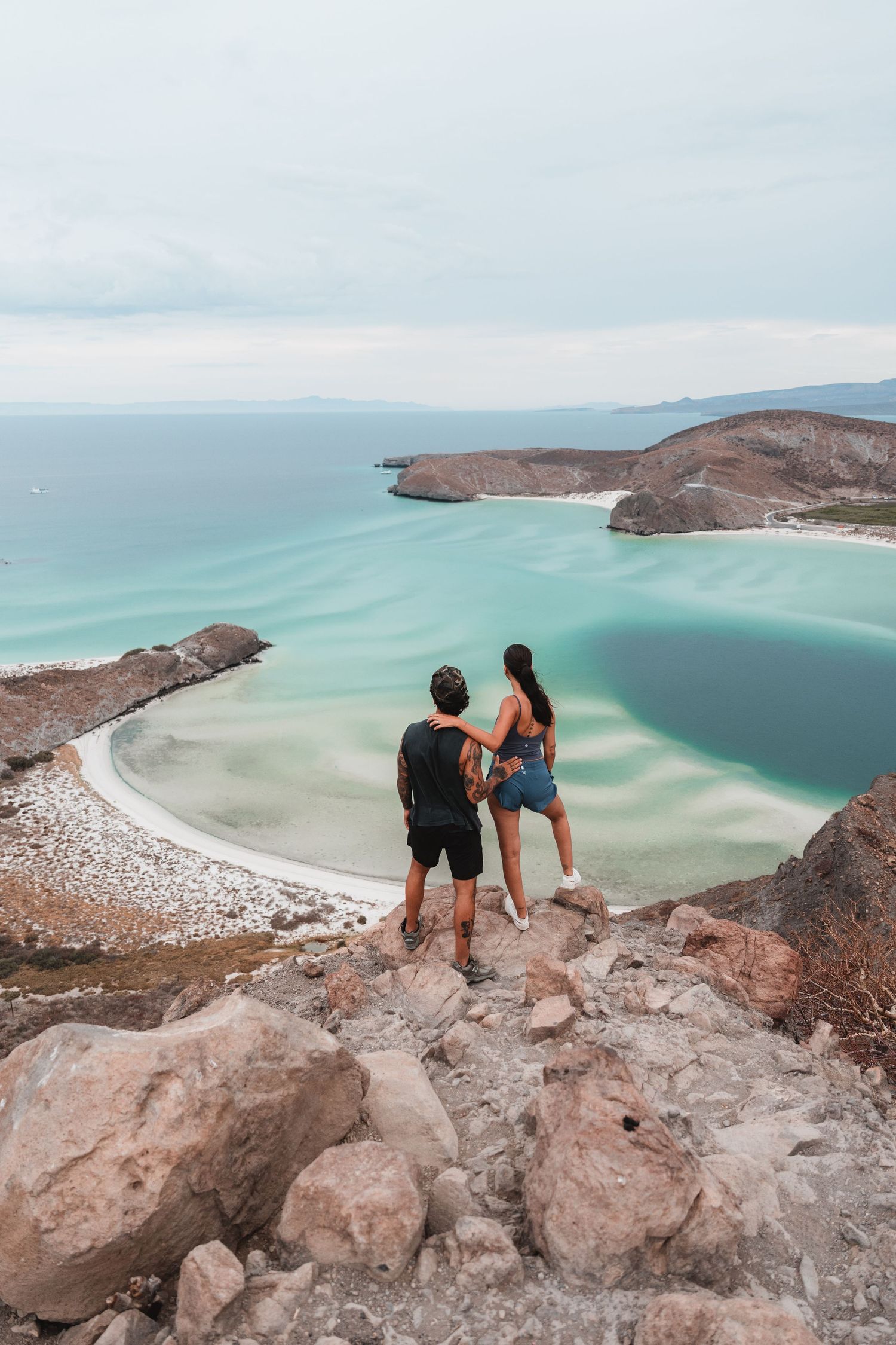 Two people standing on rocky cliff overlooking turquoise bay and coastal landscape.