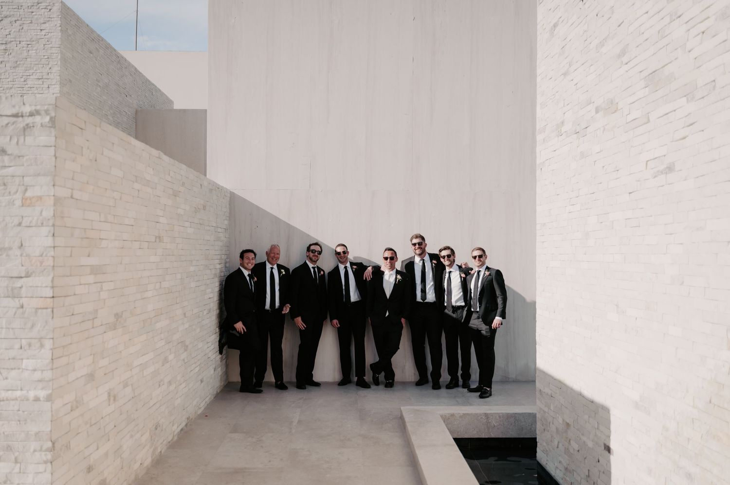 Groomsmen dressed in black suits pose together against white stone architectural backdrop.