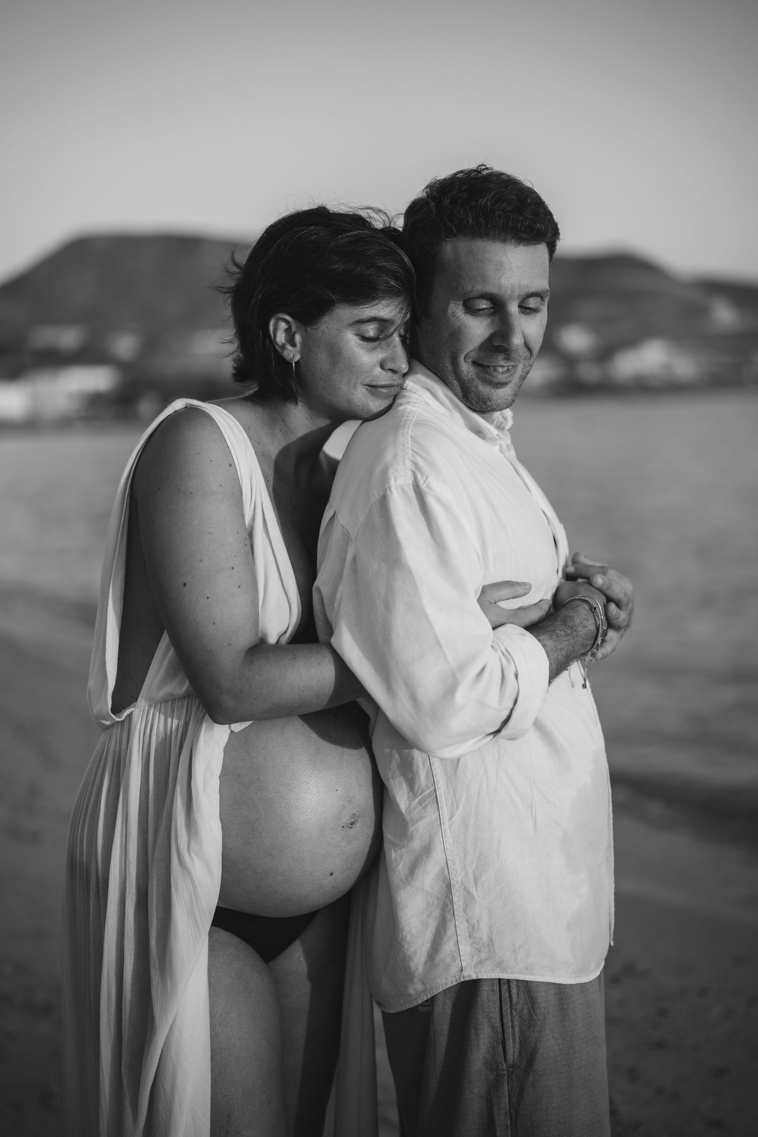 Intimate beach maternity photo of couple embracing in white clothing against coastal backdrop.