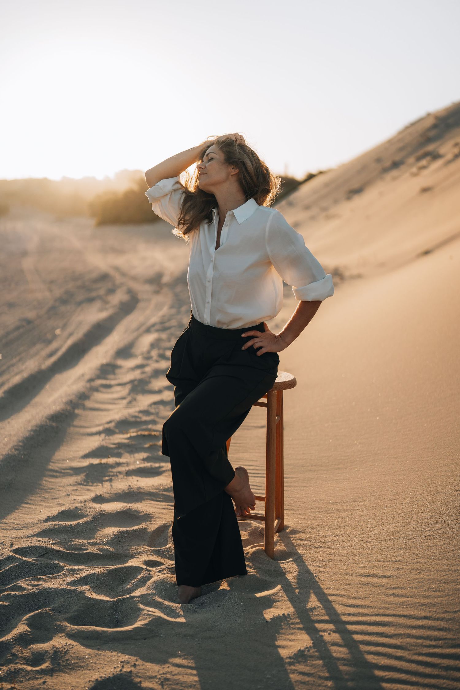 Person in white shirt and dark skirt sitting on wooden chair in sand dunes at sunset.