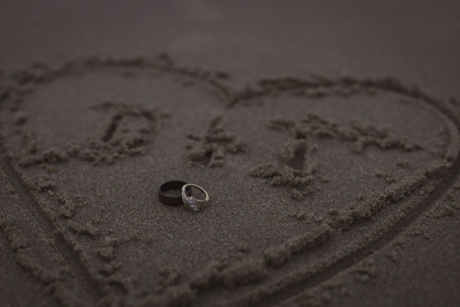 Close-up of wedding bands next to heart shape etched in sandy beach.