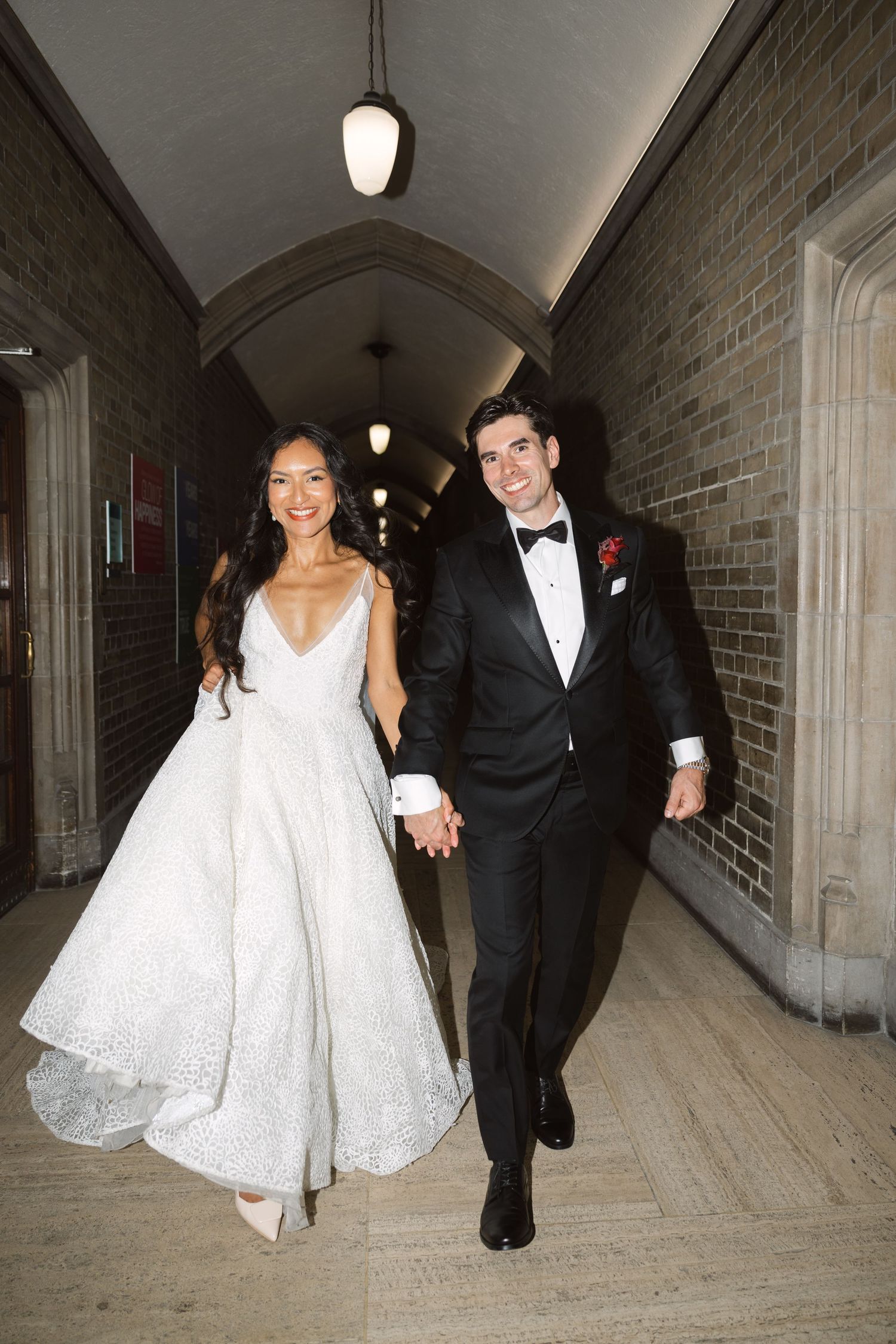 Newlyweds walk hand in hand through gothic stone corridor with dramatic lighting.