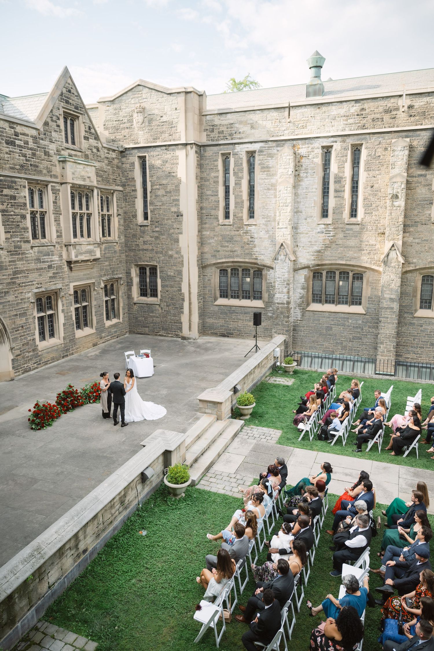 Wedding ceremony setup in a stone courtyard with white chairs and gothic architecture.