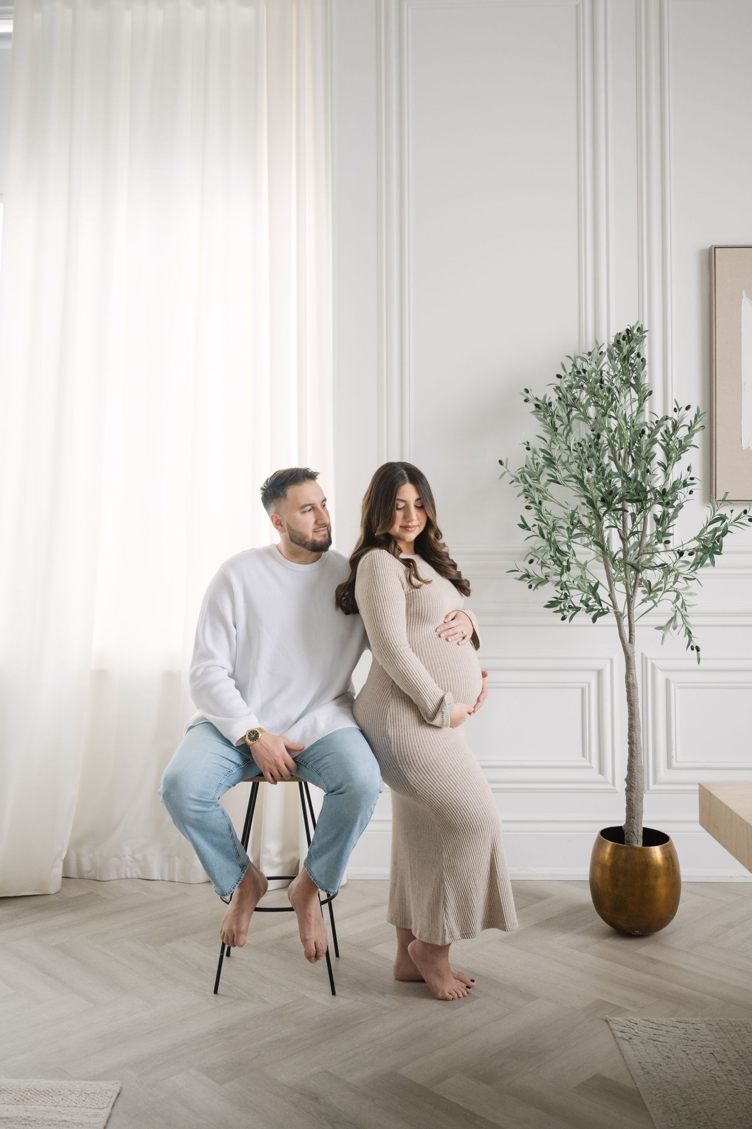 Pregnant woman in beige dress with partner sitting on chair in bright white studio room with olive tree.
