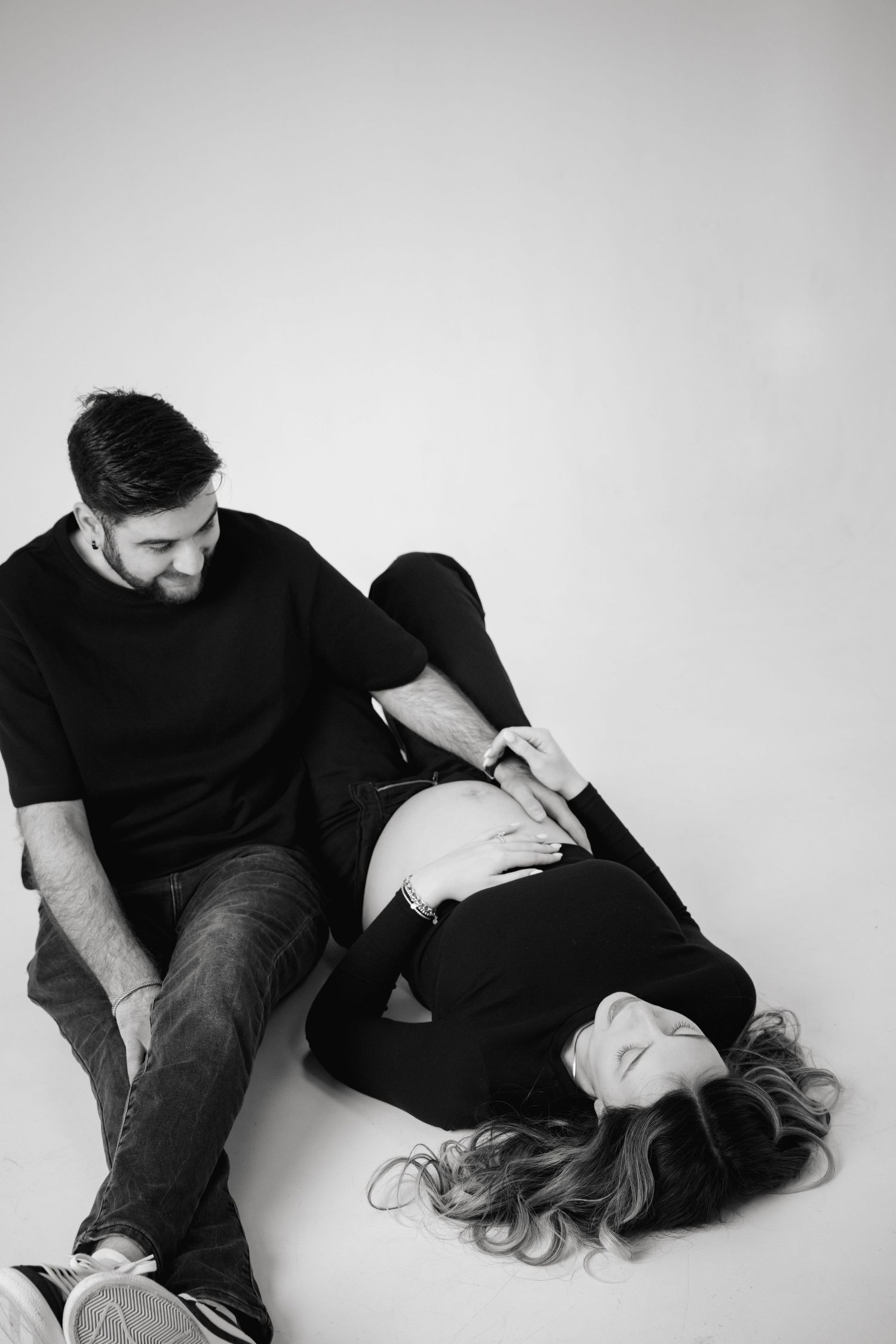 Black and white photo of a smiling man sitting beside his pregnant partner lying on the floor in a studio.