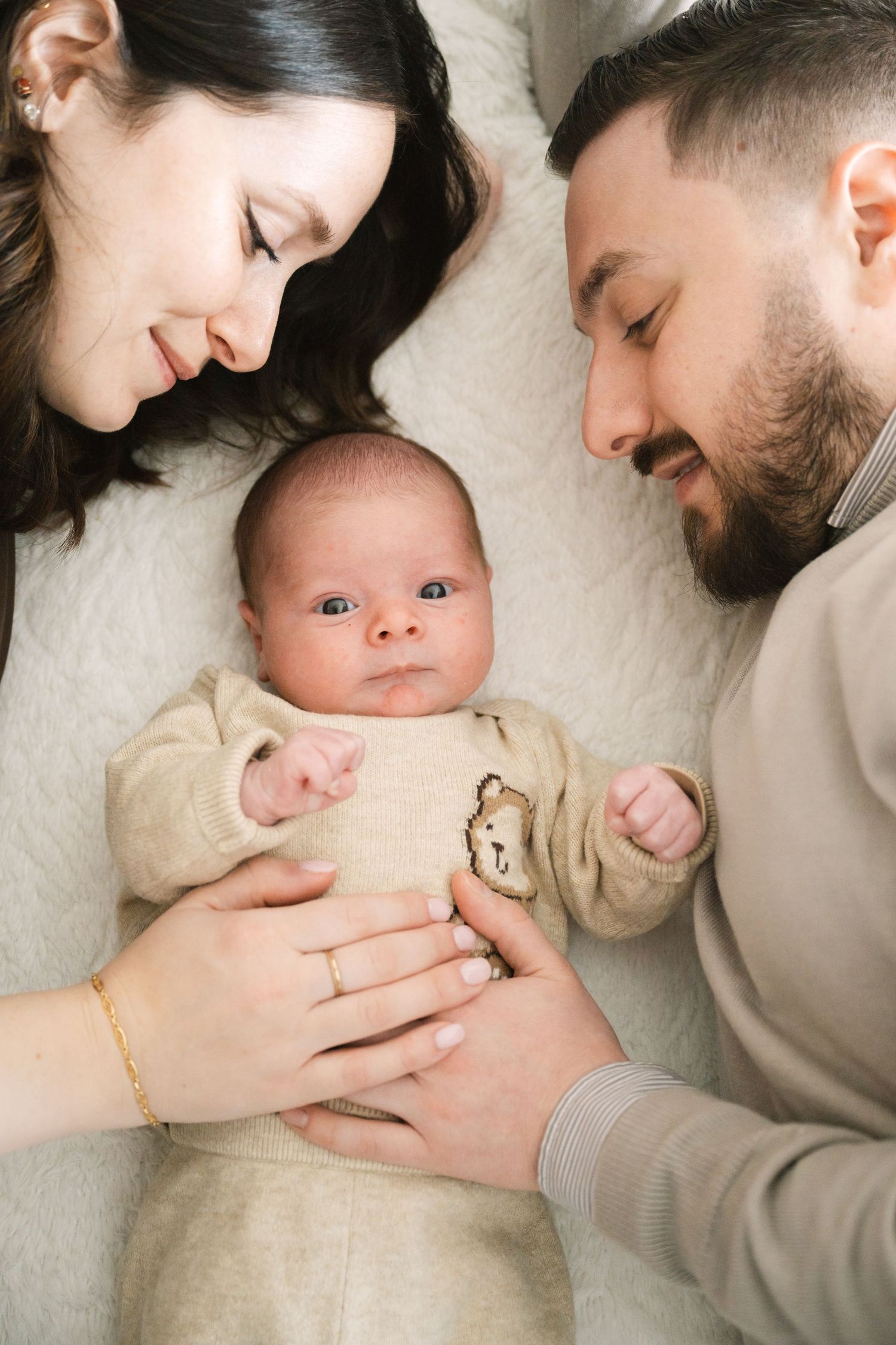 Happy parents lying with their newborn baby dressed in beige onesie on a white blanket.