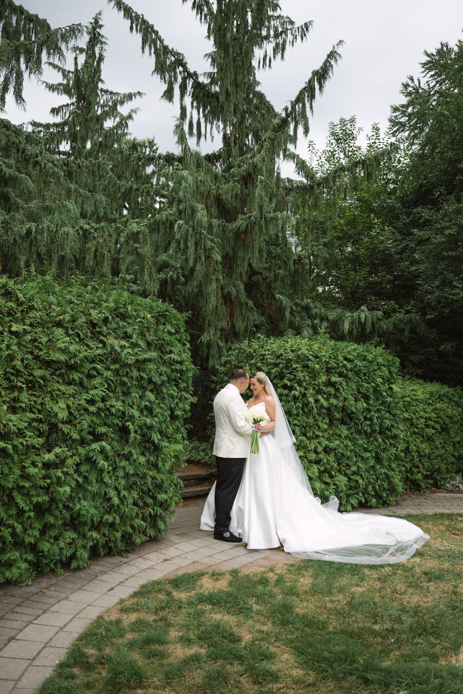 Newlyweds share an intimate moment along a garden path surrounded by evergreen trees.