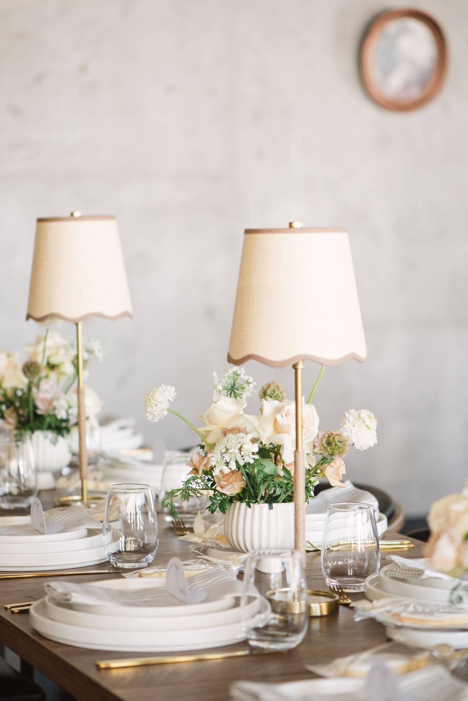 Elegant wedding table setting with scalloped cream lamps, white floral centerpieces, layered white plates, and gold accents on a wood table.