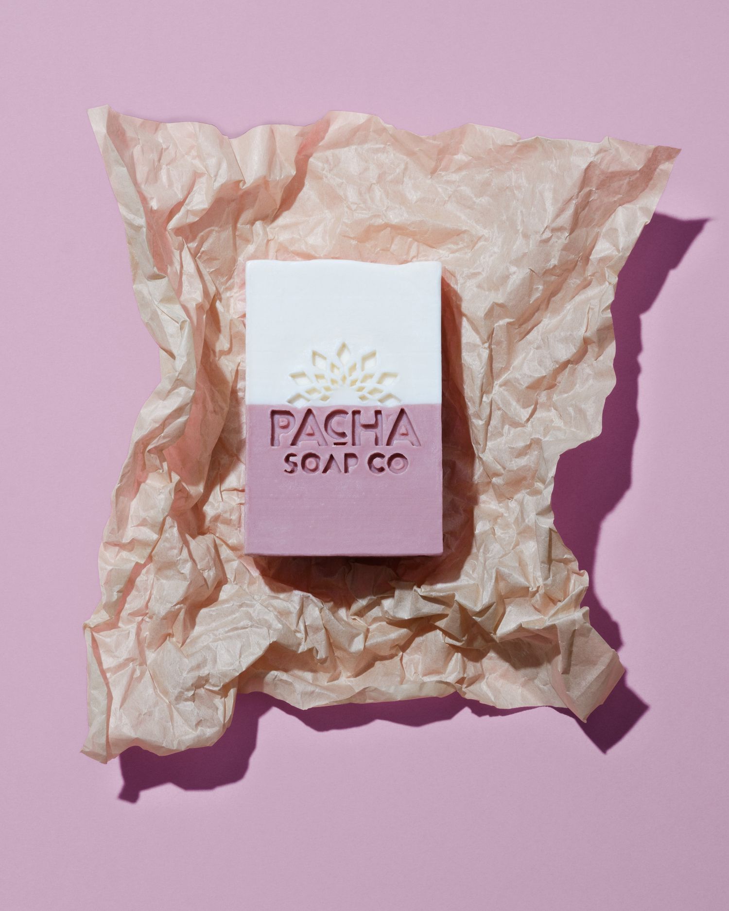 A bar of artisanal 'Pacha Soap Co' soap displayed on crumpled brown paper against a pink background.