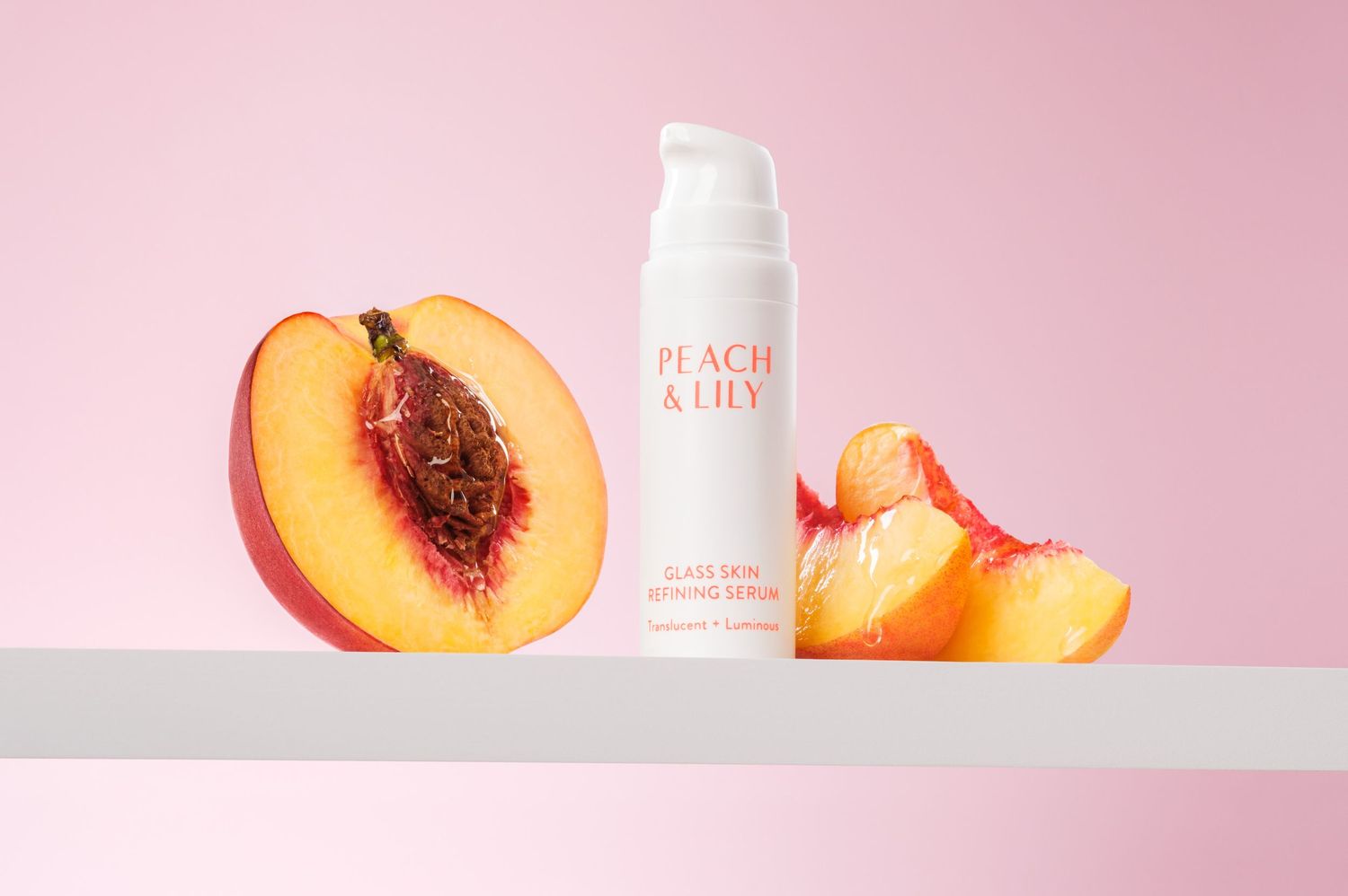Peach & Lily glass skin serum with fresh peach slices sitting on a white shelf against a bright pink background. High end studio photo.