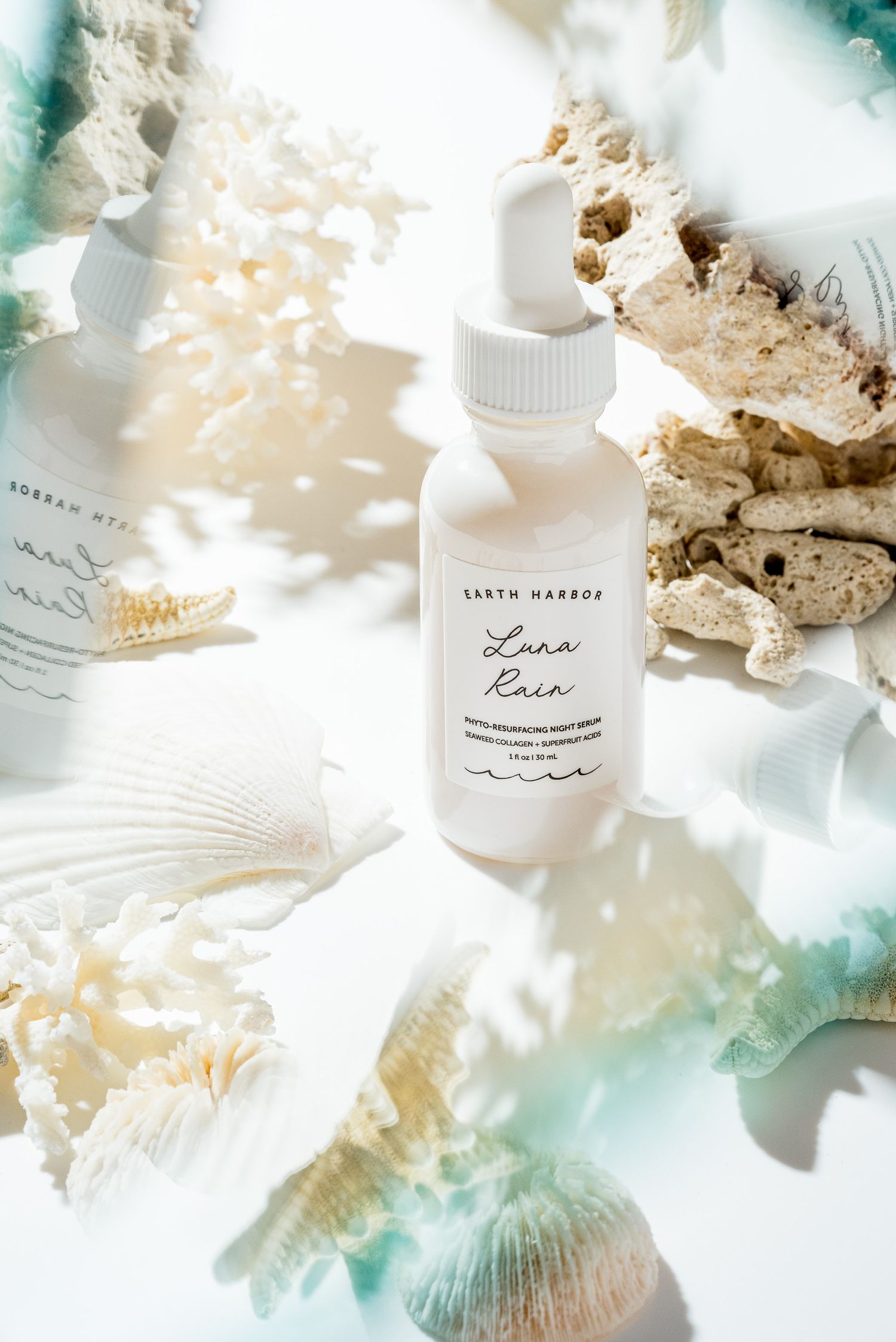 White cosmetic bottle from Earth Harbor arranged with seashells and coral on a light cream background with dreamy teal lighting effects.