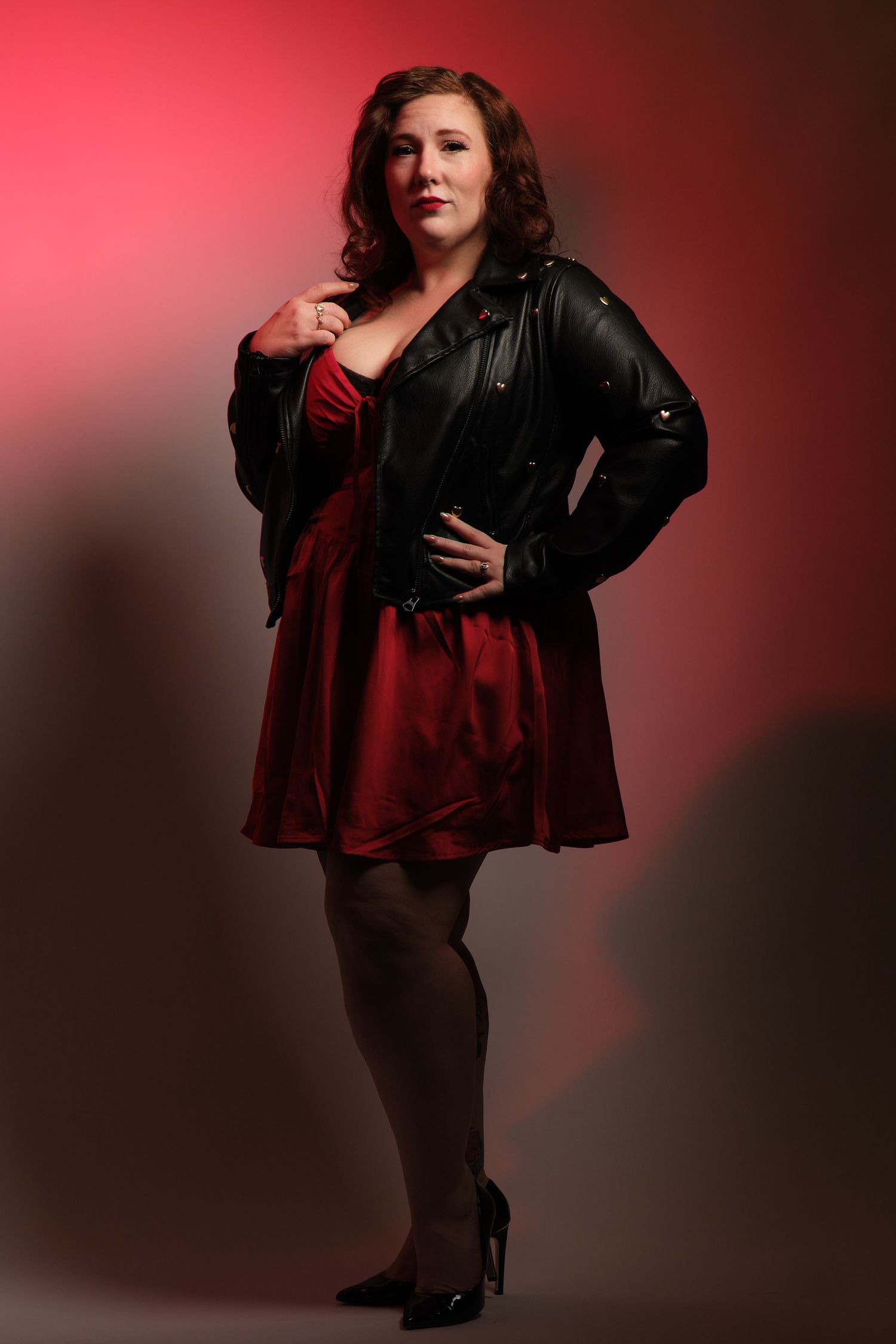 Full-length portrait of woman red satin dress, black moto jacket, and heels against colorful studio backdrop with dramatic lighting.