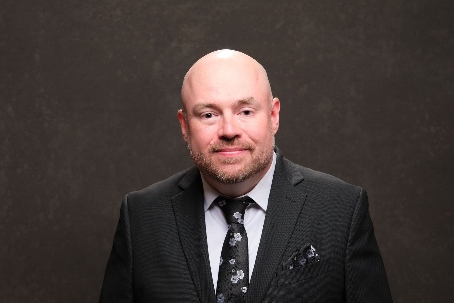 Professional headshot of man in dark gray suit with patterned tie against neutral background