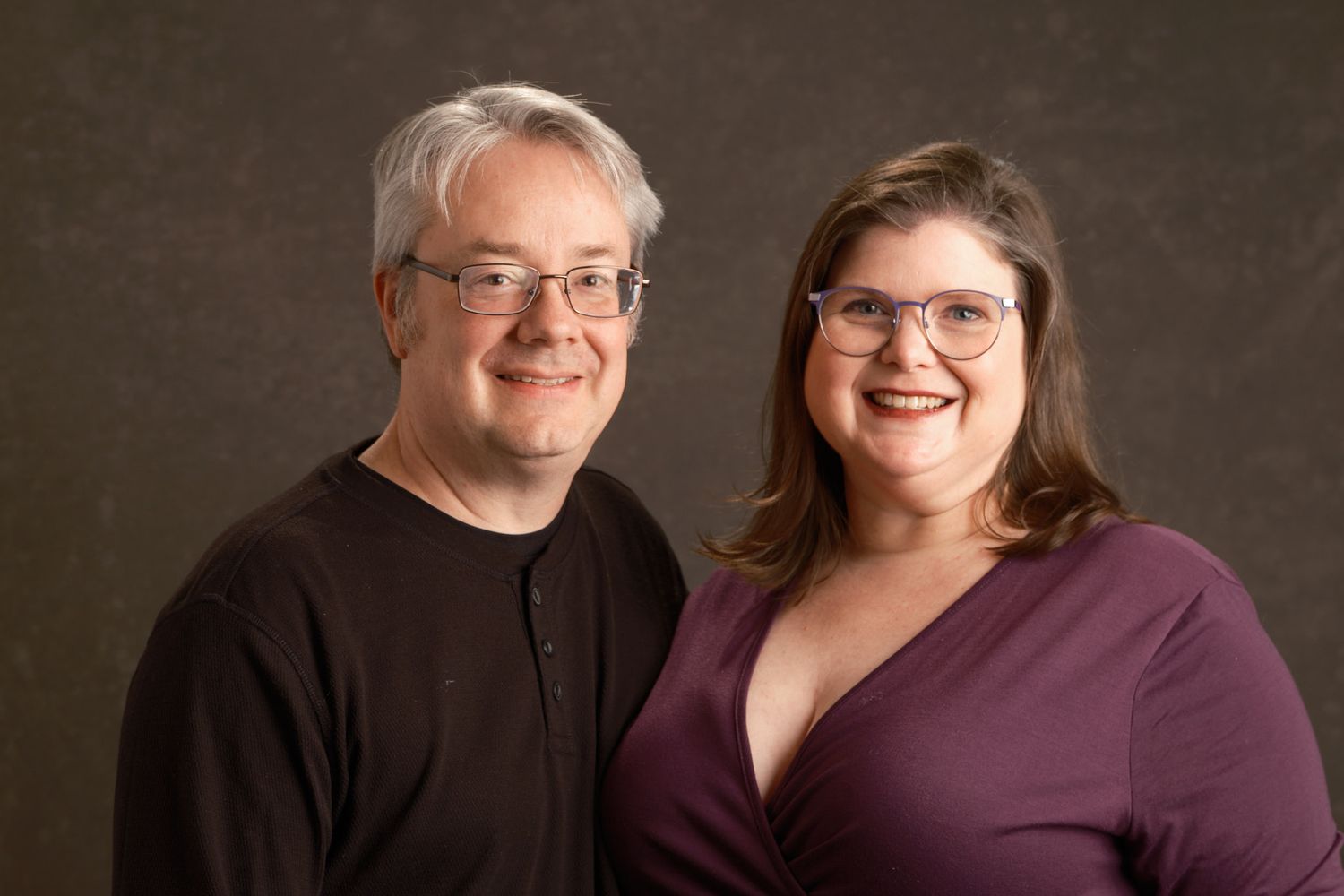 Professional couple's portrait of a man and woman smiling warmly at the camera against a neutral gray background
