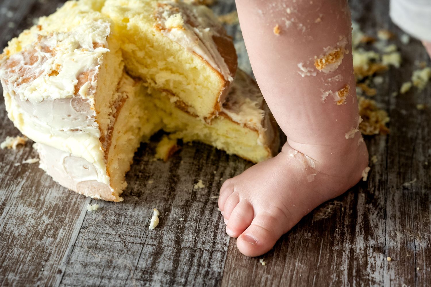 A small baby foot next to a partially eaten birthday cake on a rustic wooden surface. Smash & Splash photography.