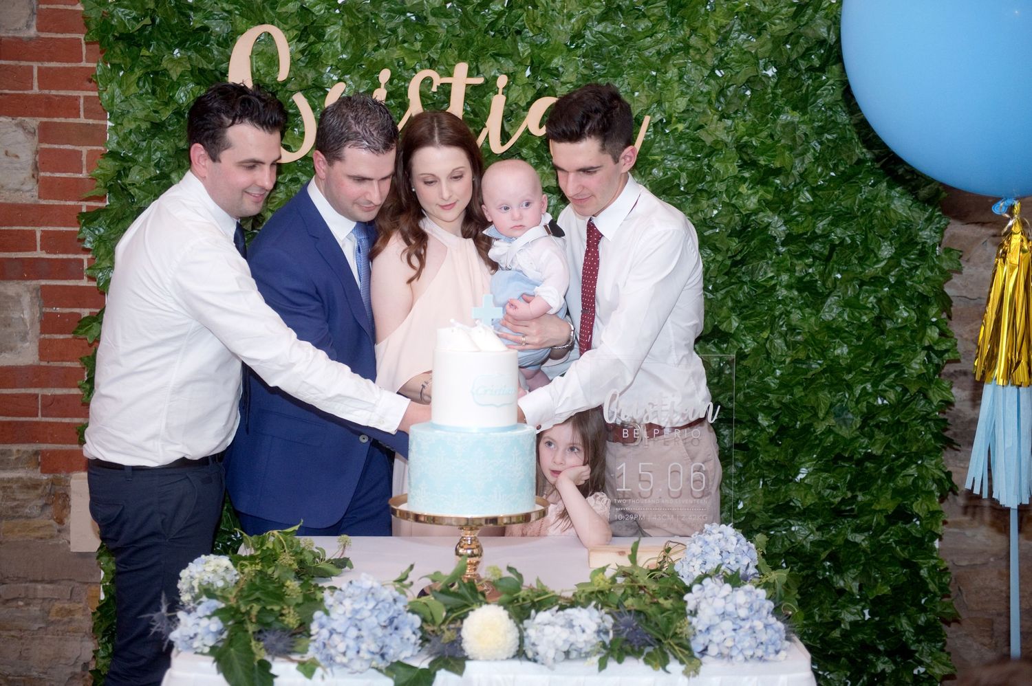 Family gathering around a blue christening cake at a baby baptism celebration with floral backdrop.