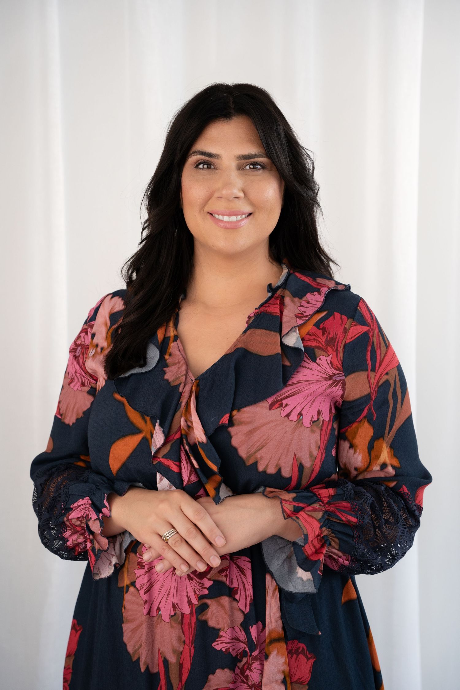 Woman in floral navy dress smiling confidently with hands clasped, posing against white backdrop.