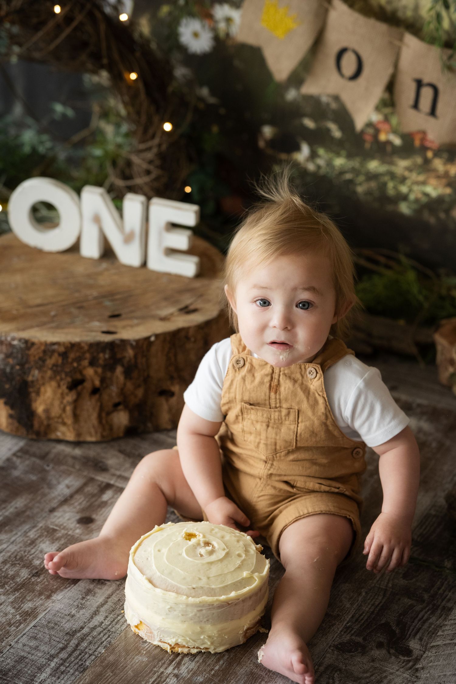 Baby in brown overalls sits beside a smash cake celebrating their first birthday with ONE letters and rustic decor.