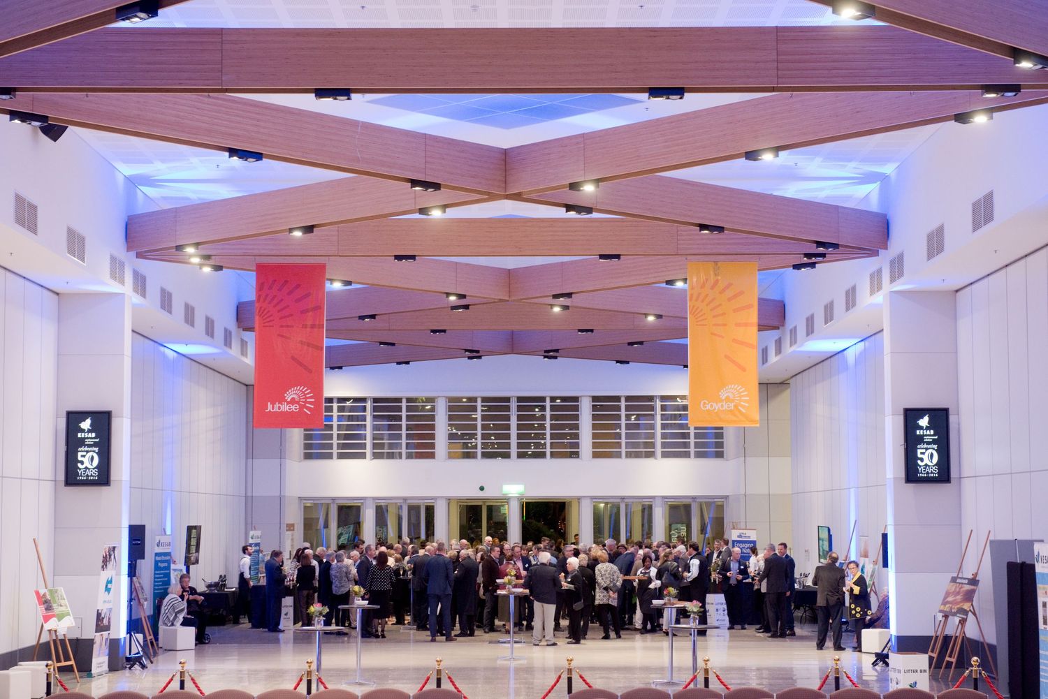 Large crowd gathering in modern lobby with purple ceiling lighting and colorful banners hanging from above. Corporate event photography.