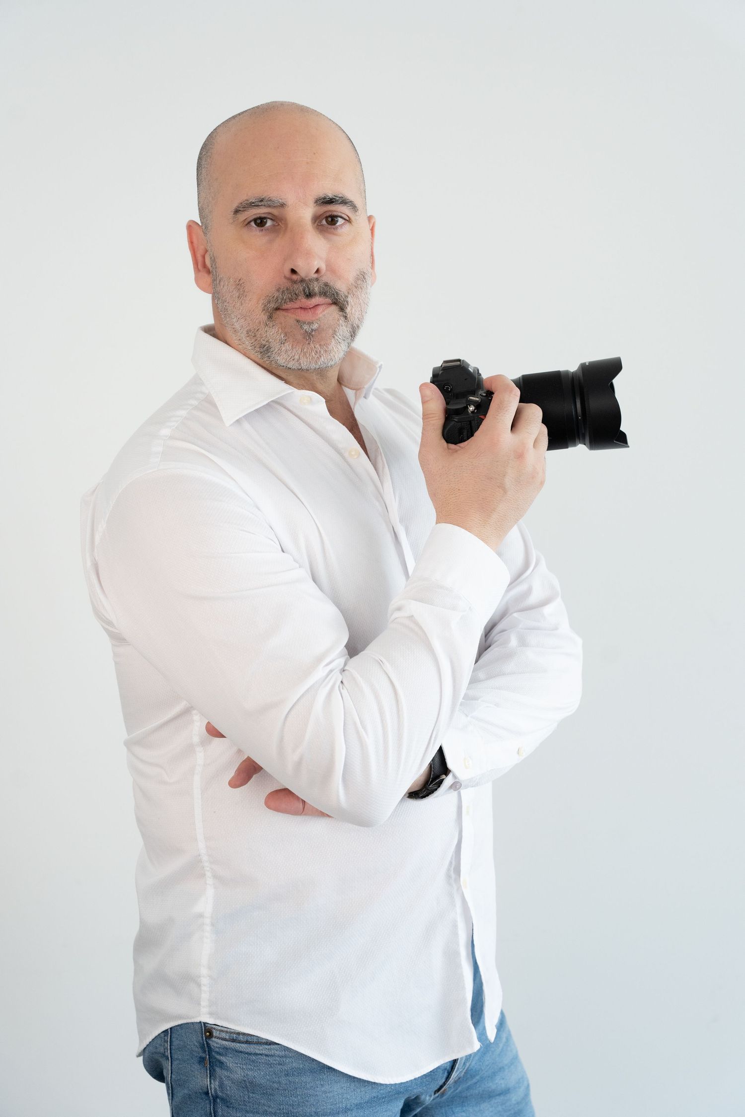 Bald man with beard in white shirt holding a black camera, posing against a white background.