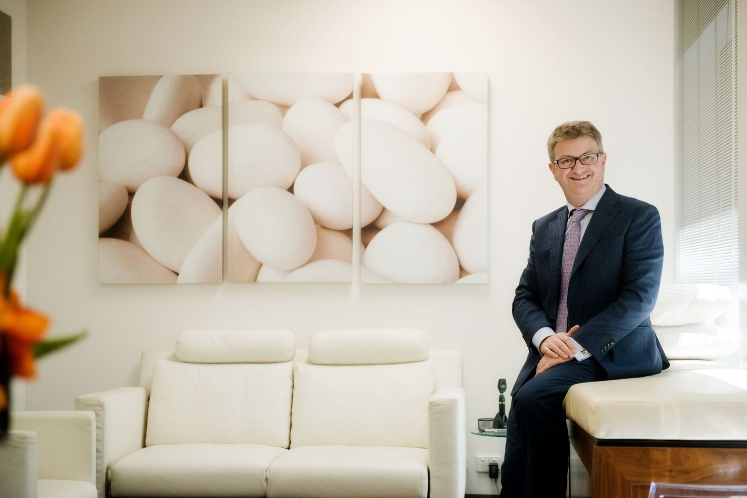 Doctor in formal attire sits comfortably on modern white sofa in bright contemporary office setting. Corporate Headshot photography.
