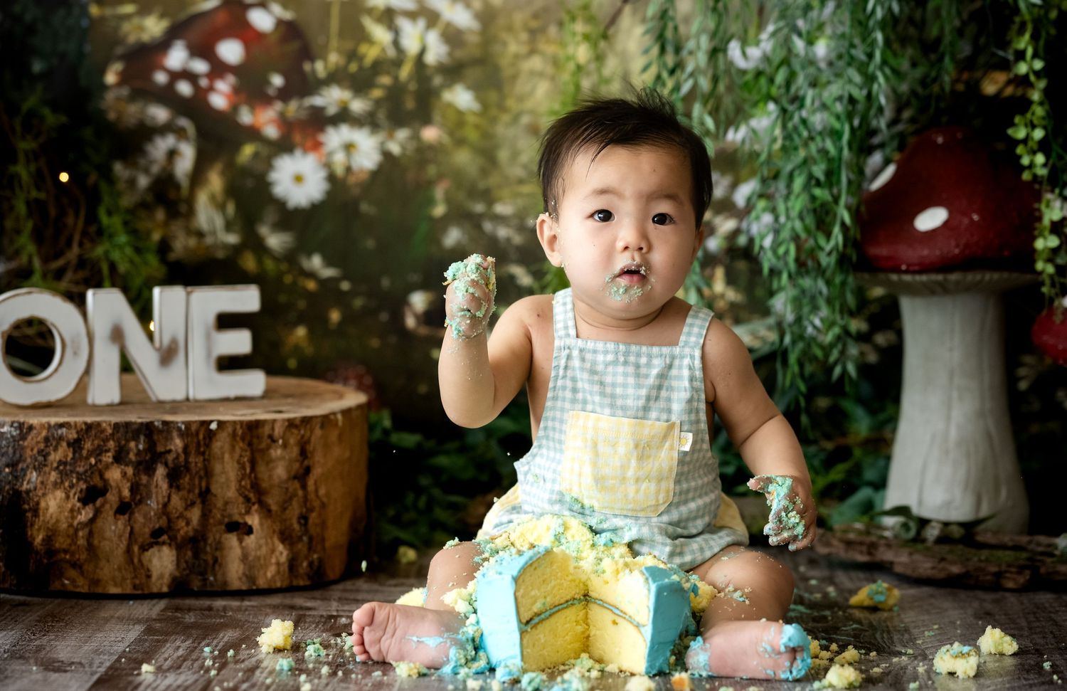 Baby boy in overalls smashing a yellow cake during a whimsical ONE birthday photoshoot with mushroom decor.