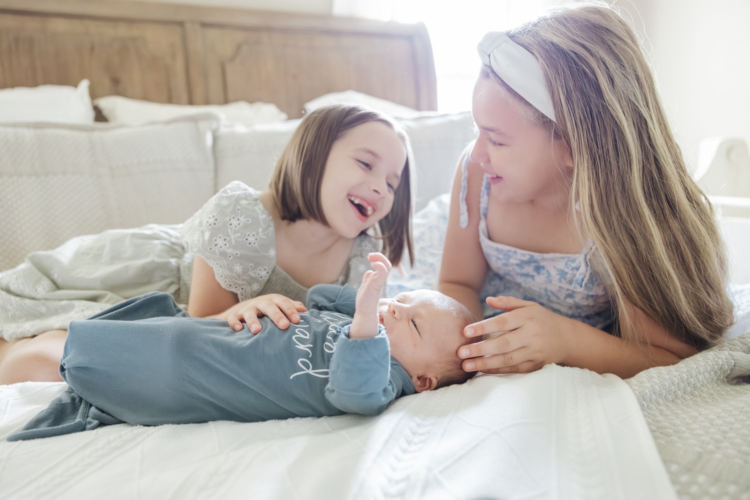 Two young sisters smile and interact with their newborn baby brother on a white bed indoors.