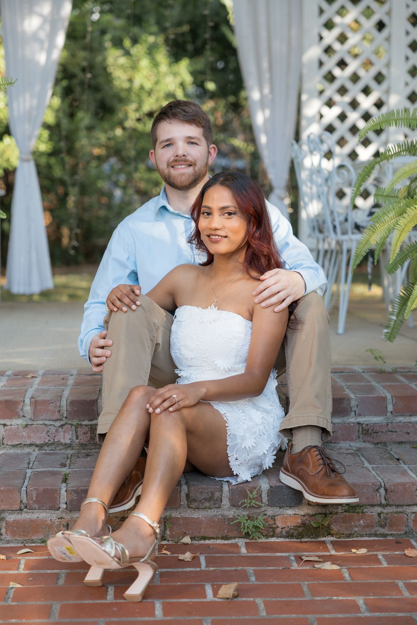 Couple sitting on brick steps, man in blue shirt embracing woman in white dress, posing in a garden setting.